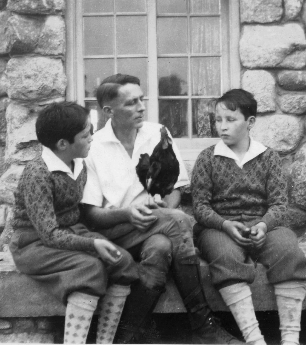 A black-and-white photo of a man and two boys sitting outside in front of a stone wall and window. The man is holding a rooster. The boys are wearing patterned sweaters and shorts, and are holding objects in their hands. They are engaged in a conversation.