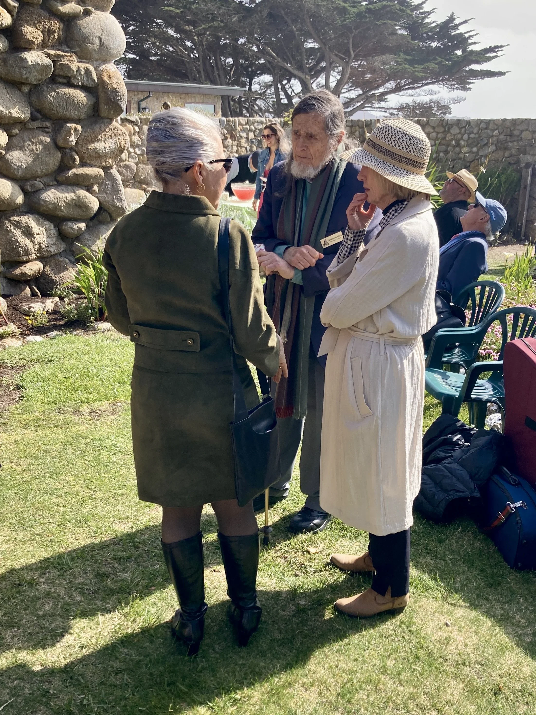 Four elderly people engaged in conversation outdoors in a garden with a stone wall and trees in the background. Two women with stylish clothing and a man with a beard and scarf are actively talking, while two other people sit in chairs nearby.