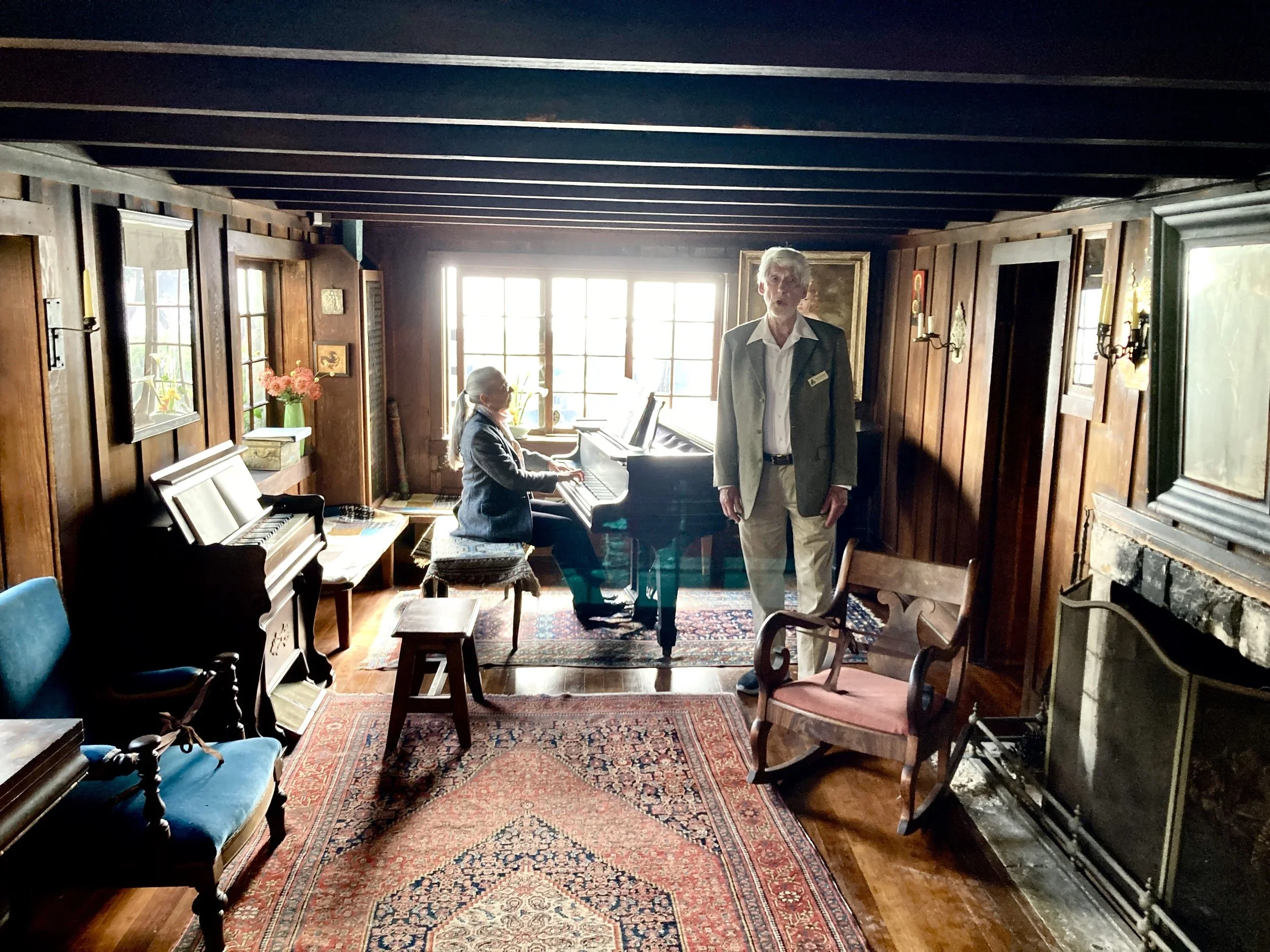 A woman playing the piano and an older man standing in a cozy, wood-paneled room with antique furniture and a fireplace.