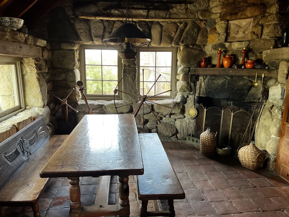 A wooden dining table with benches, large stone fireplace with a wooden mantel displaying orange pots and decorations.