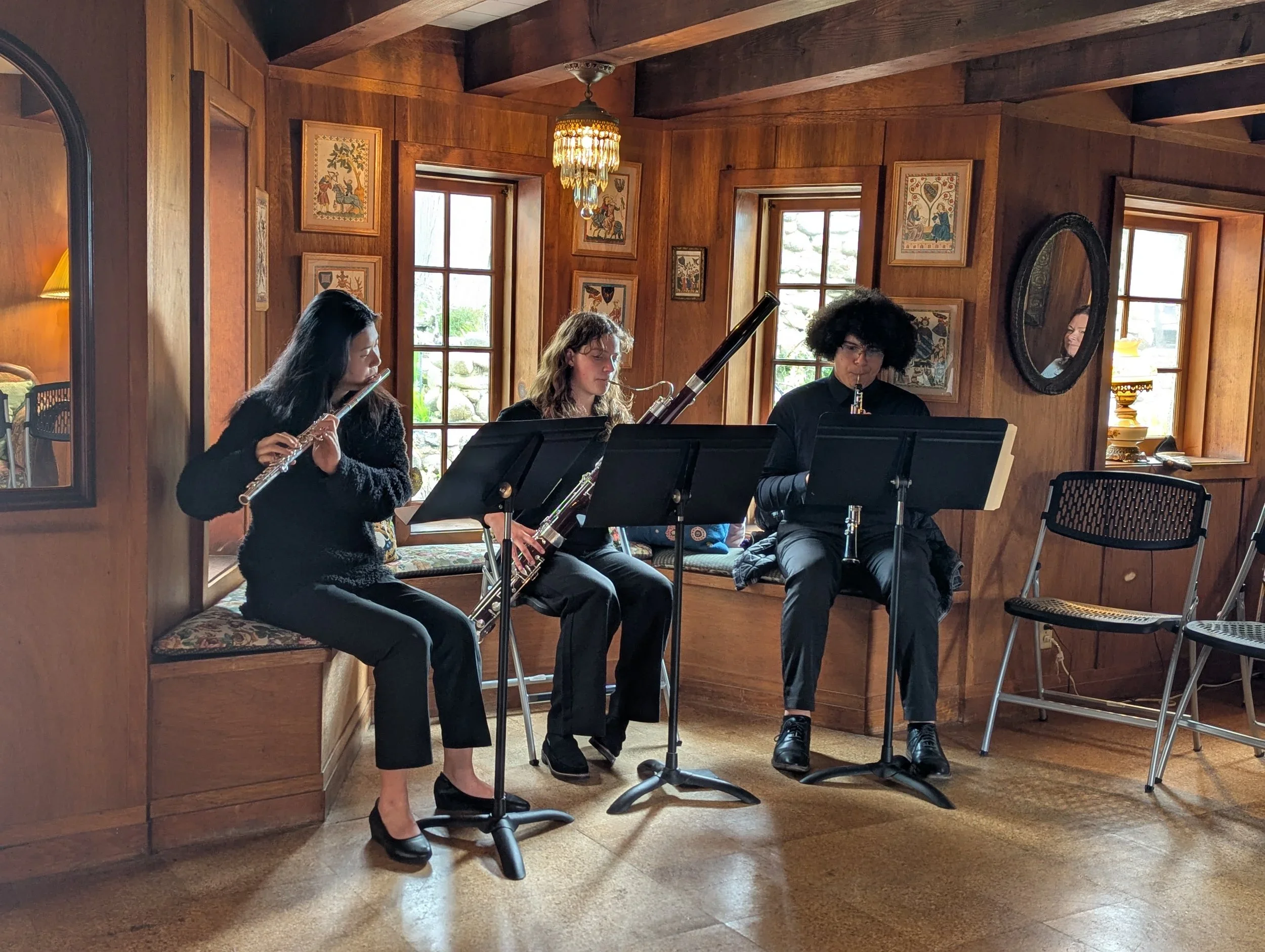 Three musicians playing woodwind instruments in a warmly lit wooden room with windows, framed artwork, and a mirror.