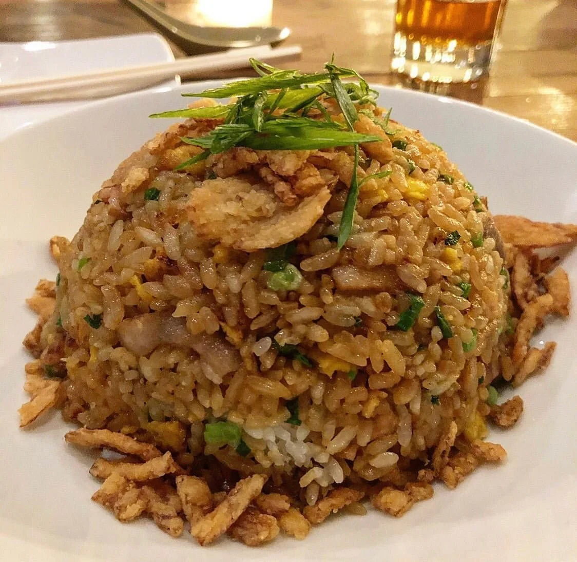 A plate of fried rice garnished with sliced green onions and shredded fried egg, presented on a white dish with a glass of beverage and chopsticks in the background.