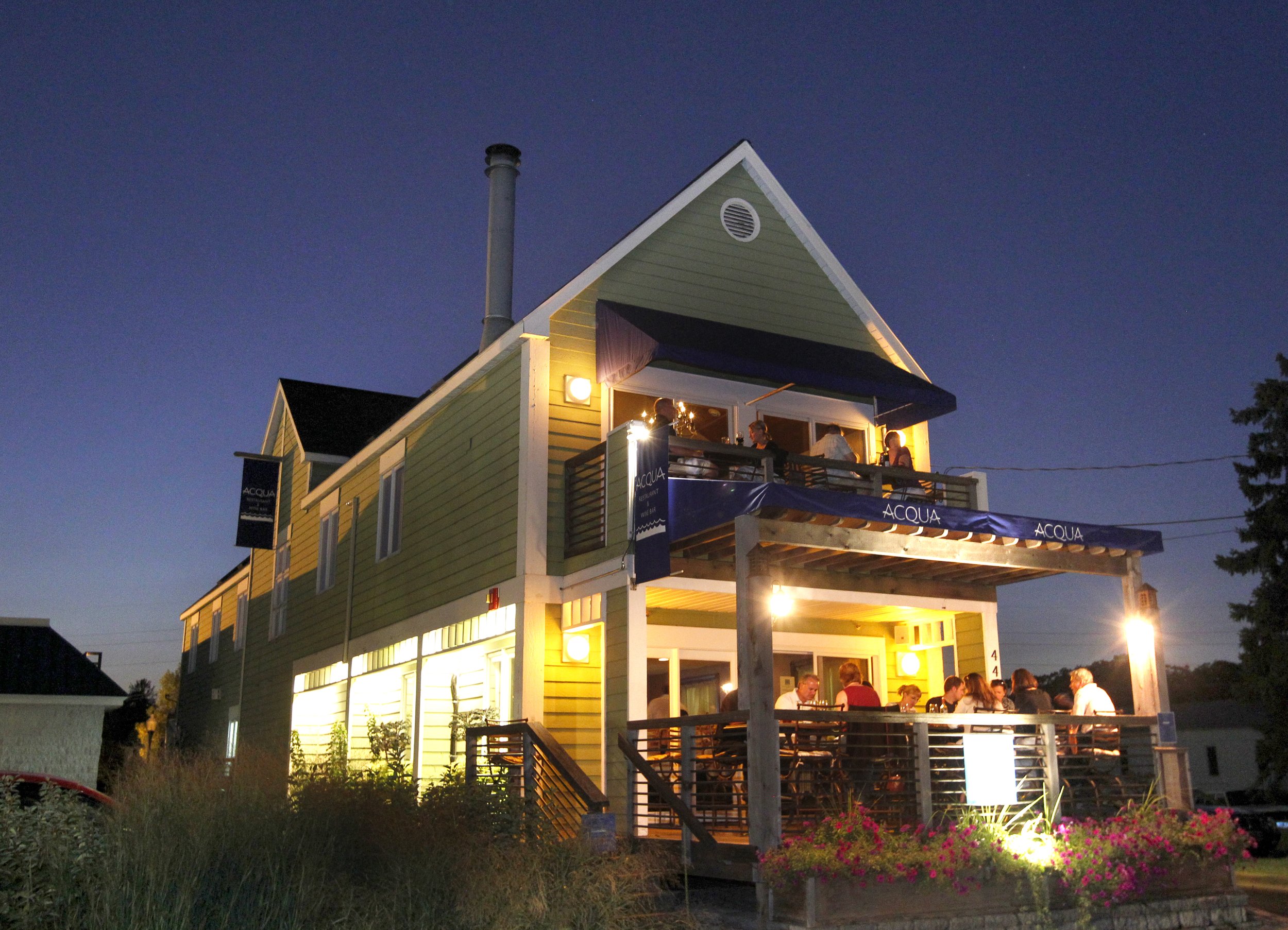A two-story house with a large patio on the upper level where people are dining at night. The house is painted yellow with white trim. The lower level has a lit outdoor area with more people dining. There are flower beds and trees around the house, and the sky is dark.