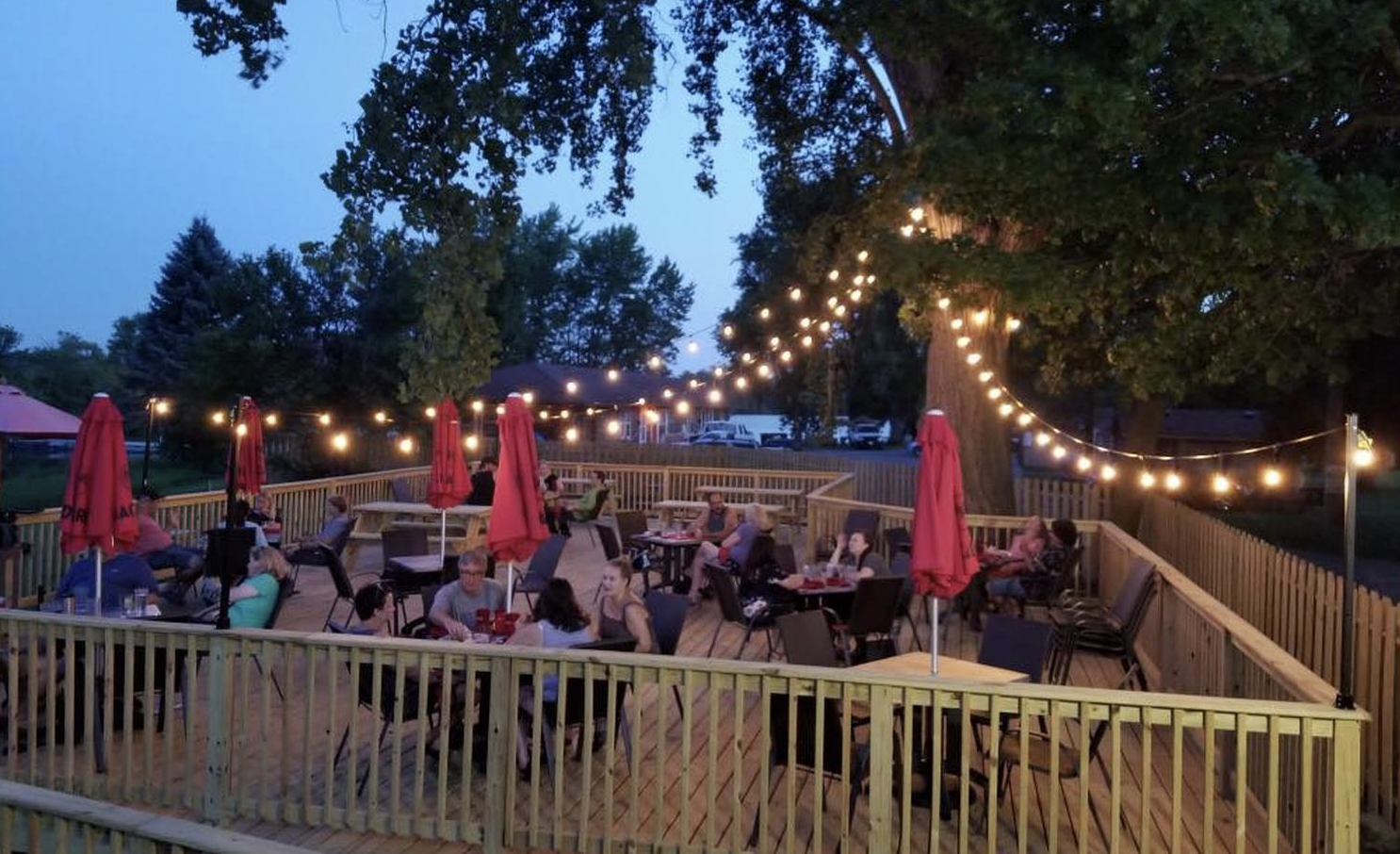Outdoor patio with wooden deck, string lights, red umbrellas, and people dining and socializing at tables during evening.