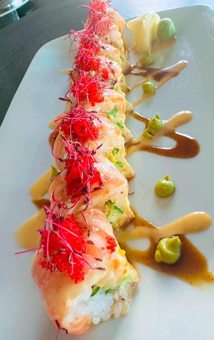 Sushi rolls topped with red fish roe and microgreens, drizzled with eel sauce, served with wasabi and pickled ginger on a rectangular white plate.