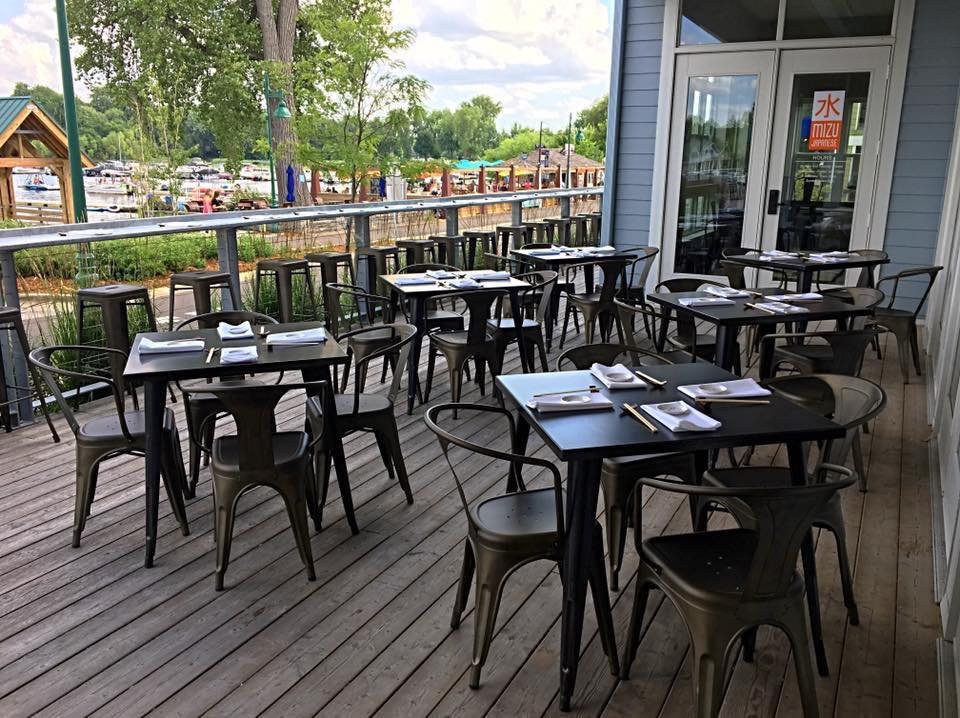 Outdoor patio of a restaurant with several black tables and chairs, each set with plates, napkins, and chopsticks. In the background, there is a view of a marina with boats and trees, and a building with large windows and a door.