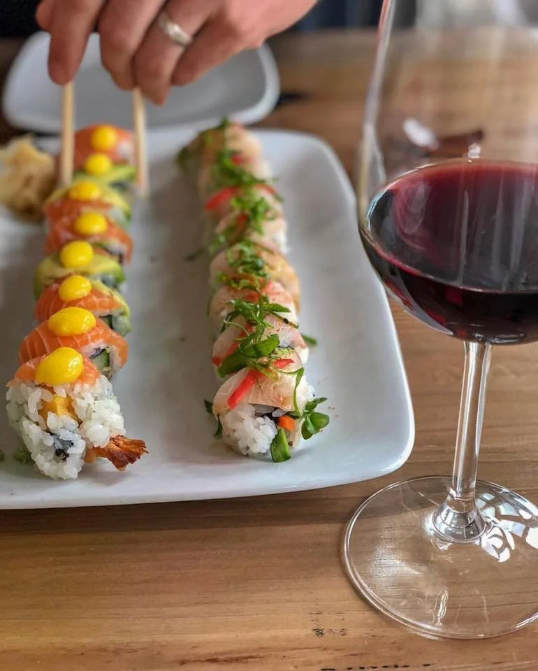 A white rectangular plate with two rolls of sushi and one hand using chopsticks to pick up sushi. A glass of red wine is on the right side of the plate on a wooden table.