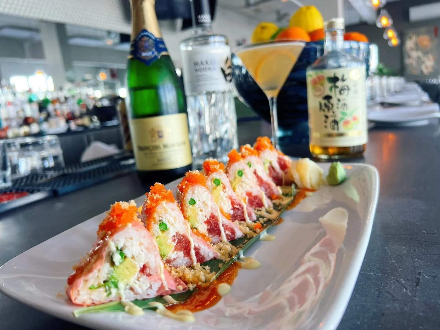 Sushi rolls with orange fish roe and green peas on a white plate, with a green wasabi dollop, served at a restaurant with a bar in the background.