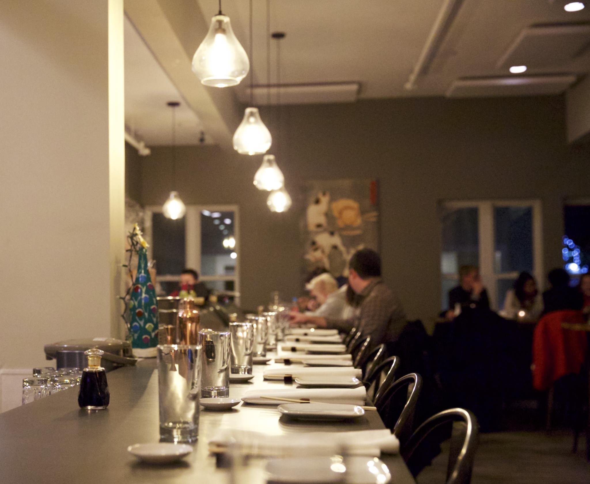 A long dining table set with glasses, plates, and rolled napkins in a restaurant with hanging pendant lights, and people seated in the background.