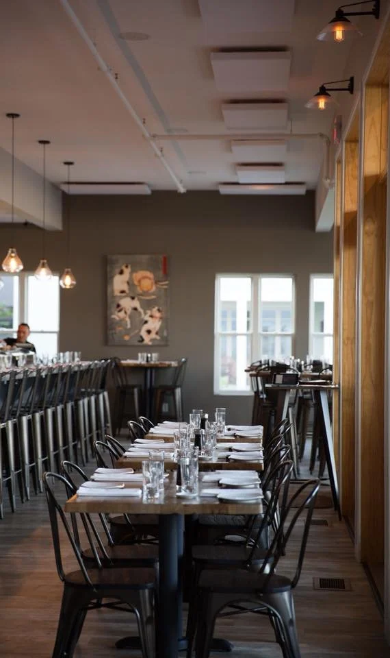 A restaurant interior with a long table set for dining, including glasses, napkins, and cutlery, with a bar area in the background and large windows providing natural light.
