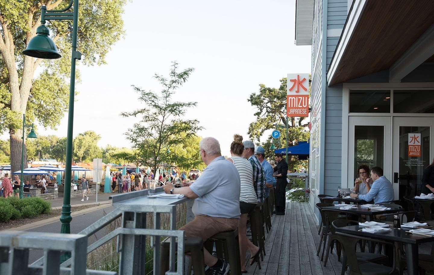 People dining on an outdoor patio of a Japanese restaurant at sunset. The patio has tables and chairs, with some people eating and others waiting. A street with trees and a crowd of people is visible in the background.