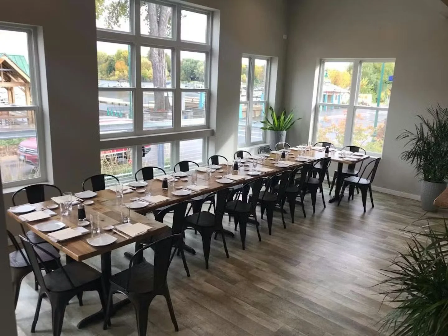 Empty restaurant dining area with two long wooden tables set with white plates, glasses, napkins, and black salt and pepper shakers. Black chairs surround the tables. Large windows on the walls let in natural light, showing outdoor trees and parked cars. Potted plants are placed near the windows.