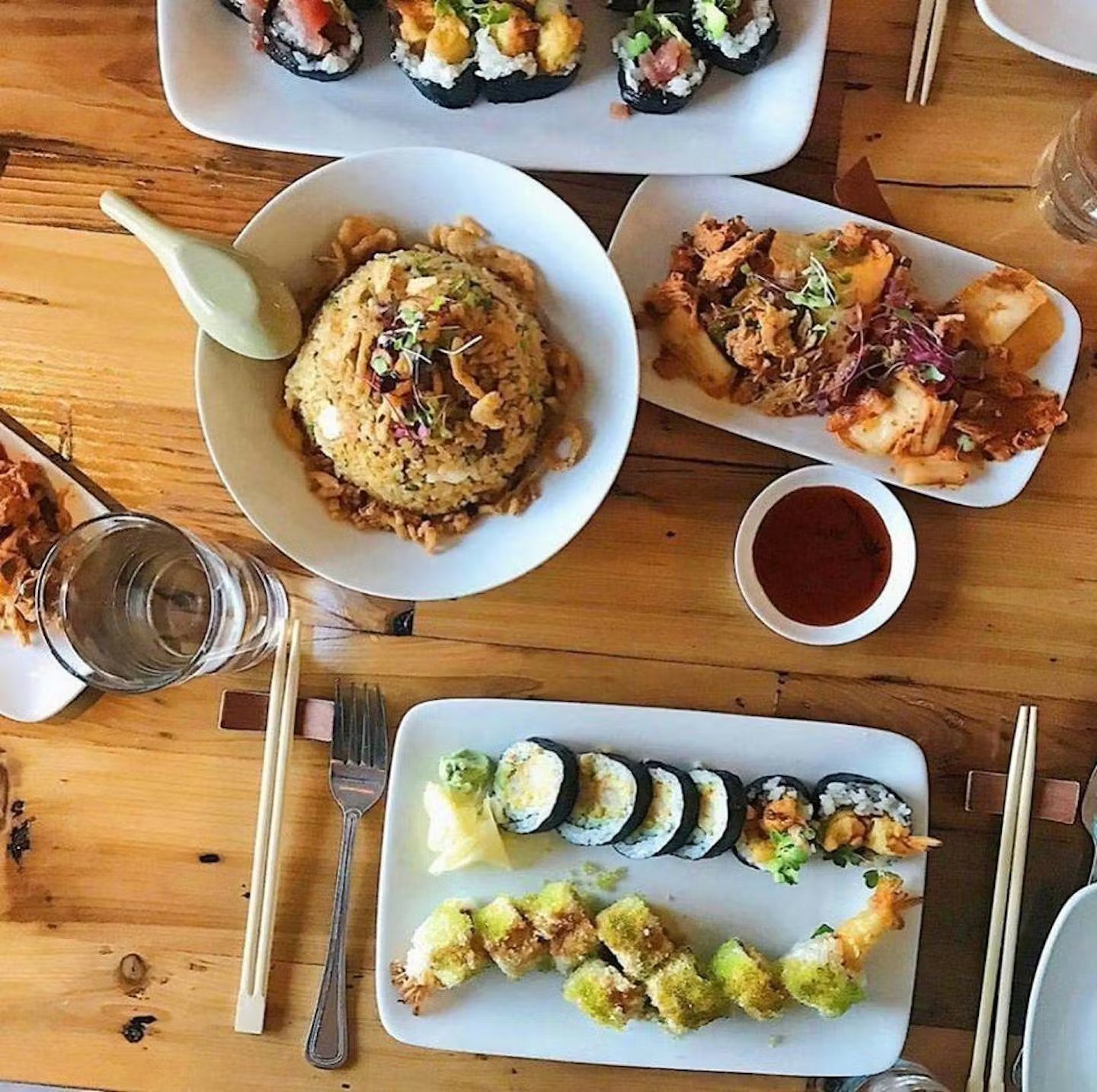 Assorted Japanese sushi dishes on white plates, including rolls with fish and vegetables, and tempura shrimp with battered vegetables, served with sauces and wasabi on a wooden table.