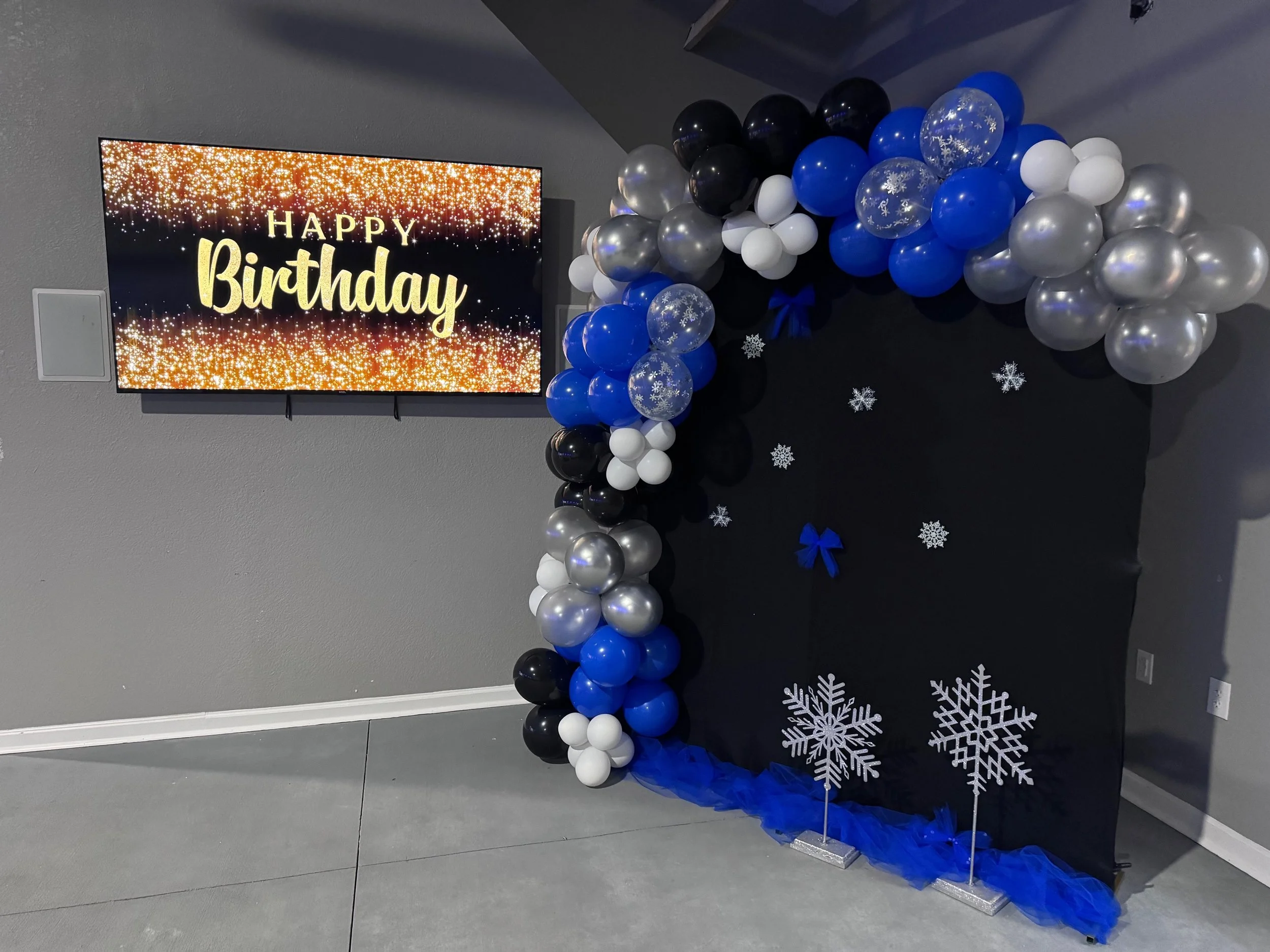 Decorative birthday photo backdrop with balloons in black, blue, silver, and white, snowflake cutouts, and a screen displaying "Happy Birthday" with a glittery background.