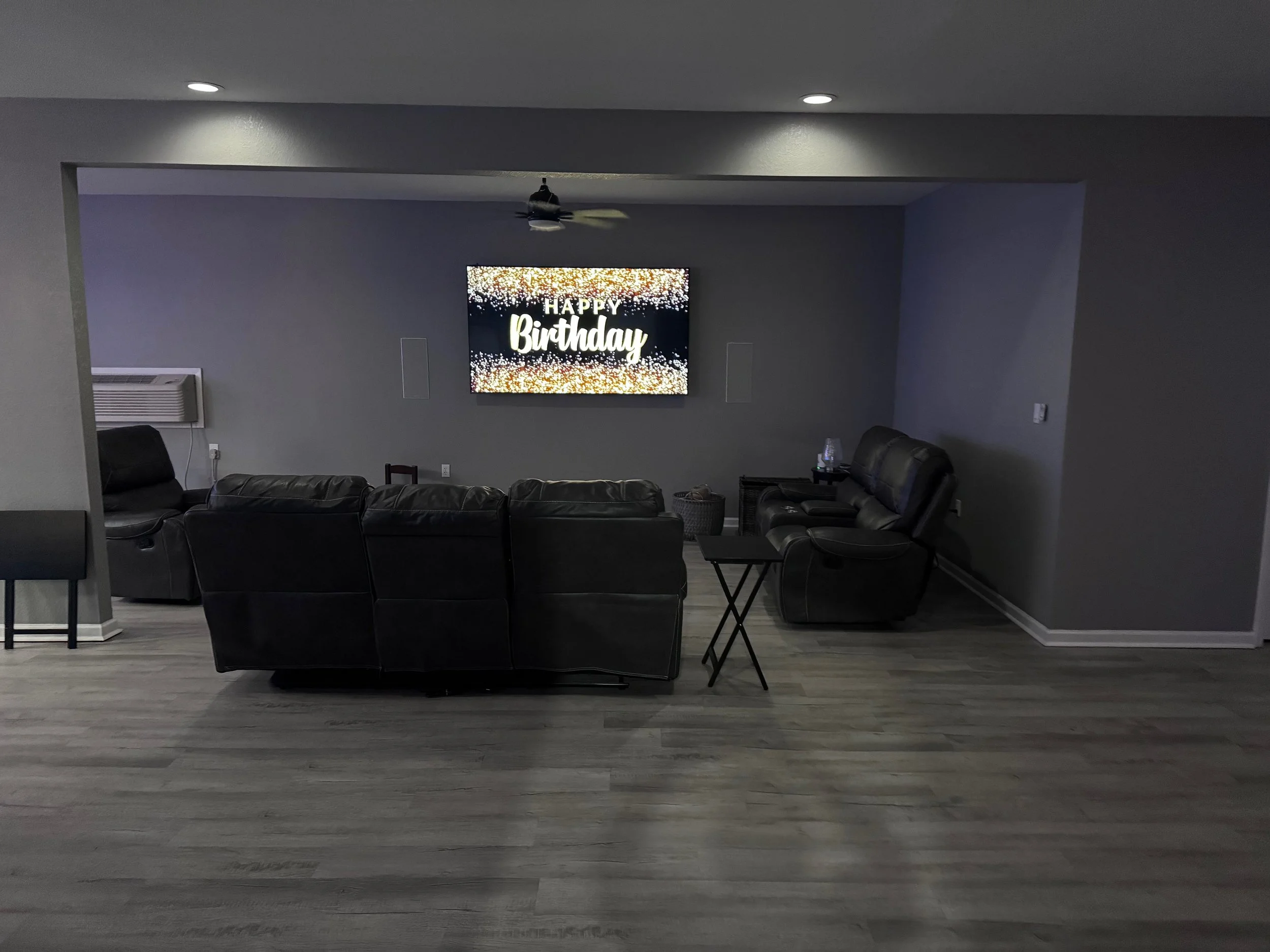 Living room with black leather couches, a small black table, a television on the wall displaying 'Happy Birthday' with a sparkly background, and a ceiling fan.