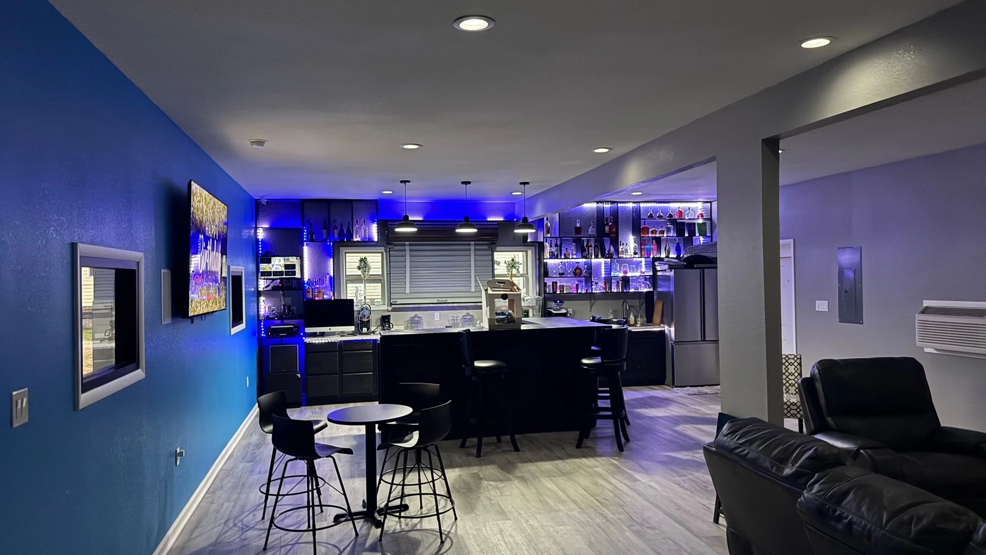 Interior of a modern basement bar with blue and purple lighting, featuring a bar counter, stools, a wall-mounted TV, and a sitting area with black leather chairs.
