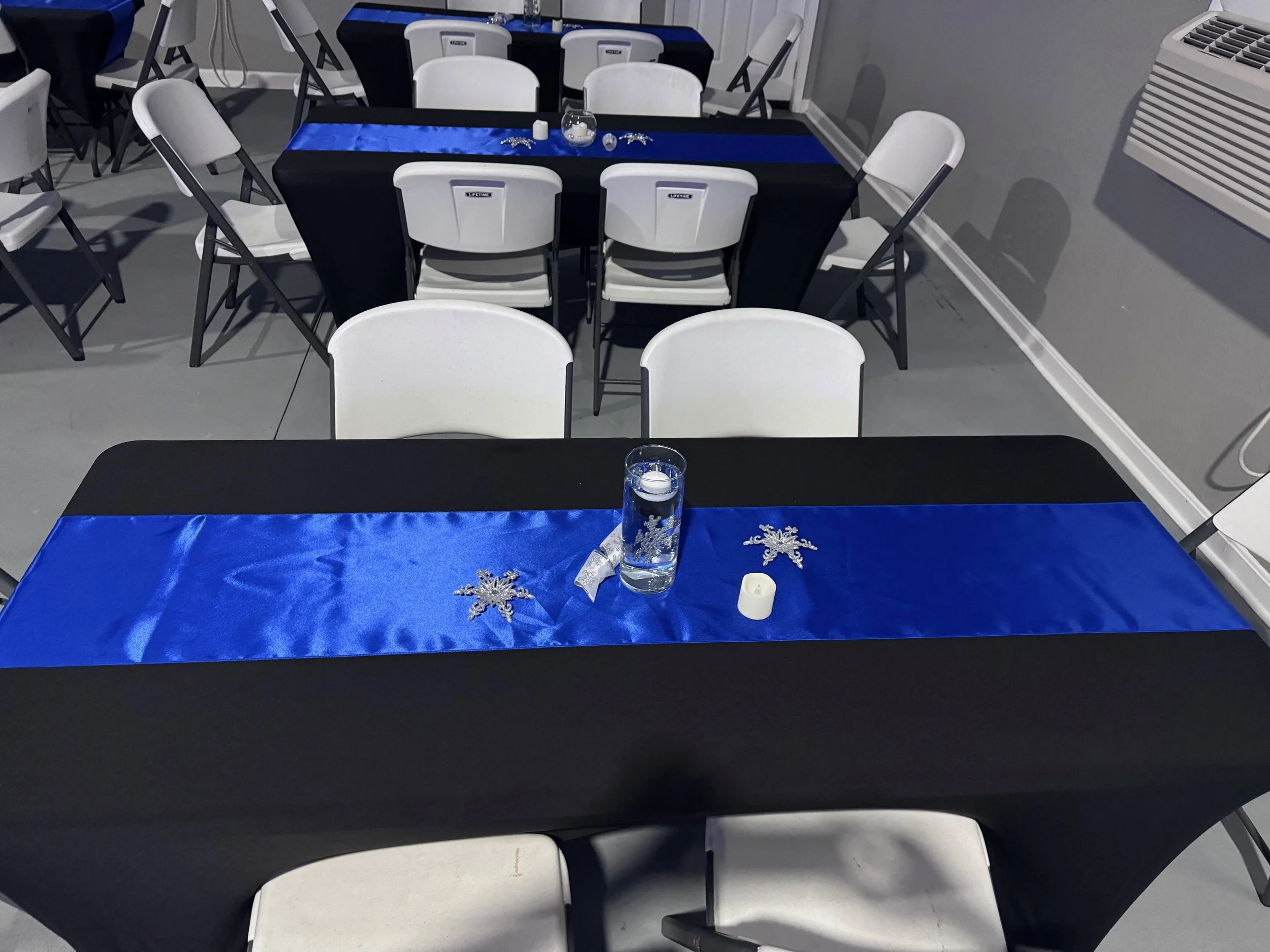Decorated tables with black tablecloths, blue satin runners, silver snowflake ornaments, a glass container with floating candles, and white chairs arranged for a festive event.