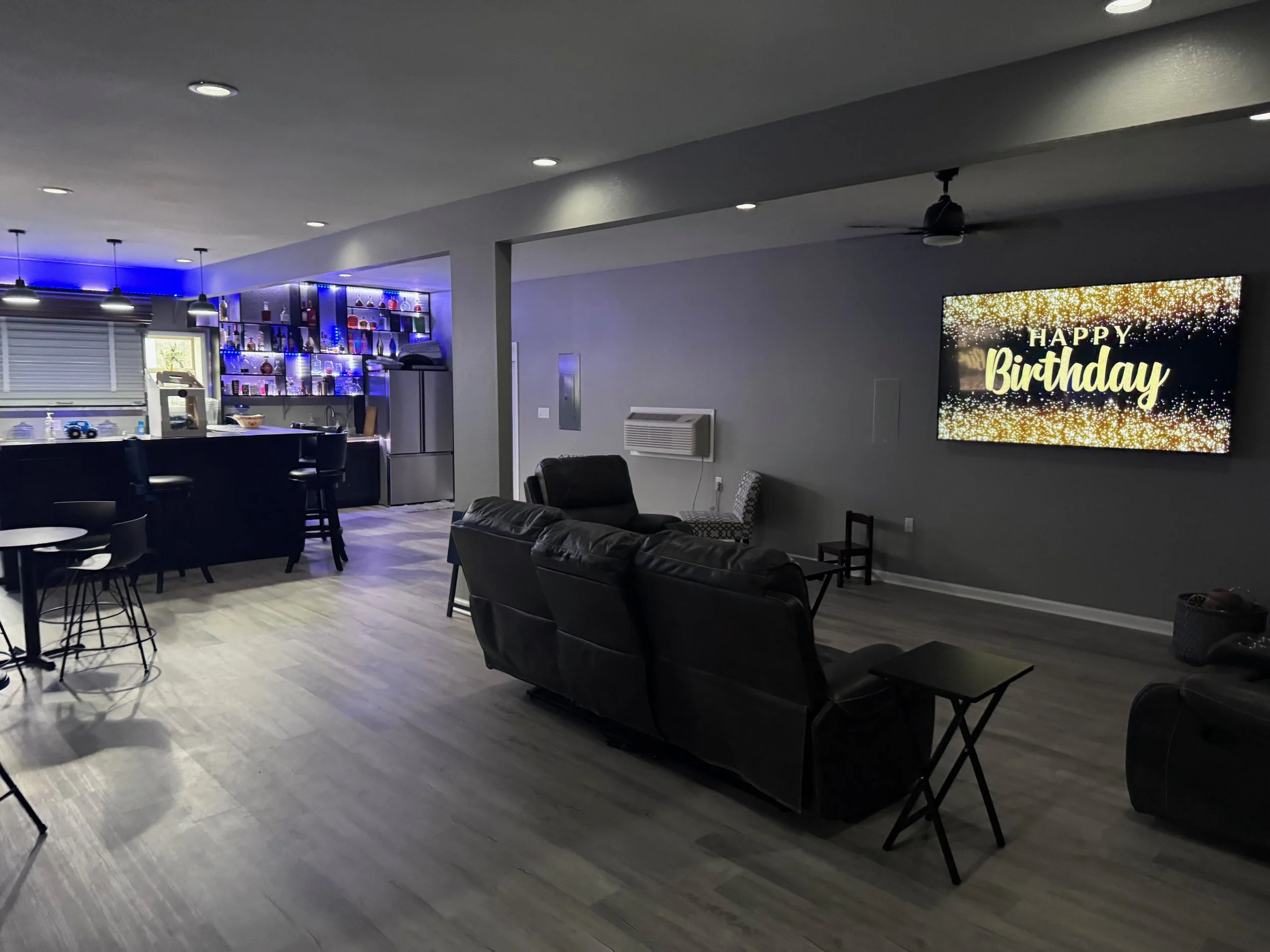 Living room with a large digital screen displaying 'Happy Birthday' and a bar area with blue lighting.