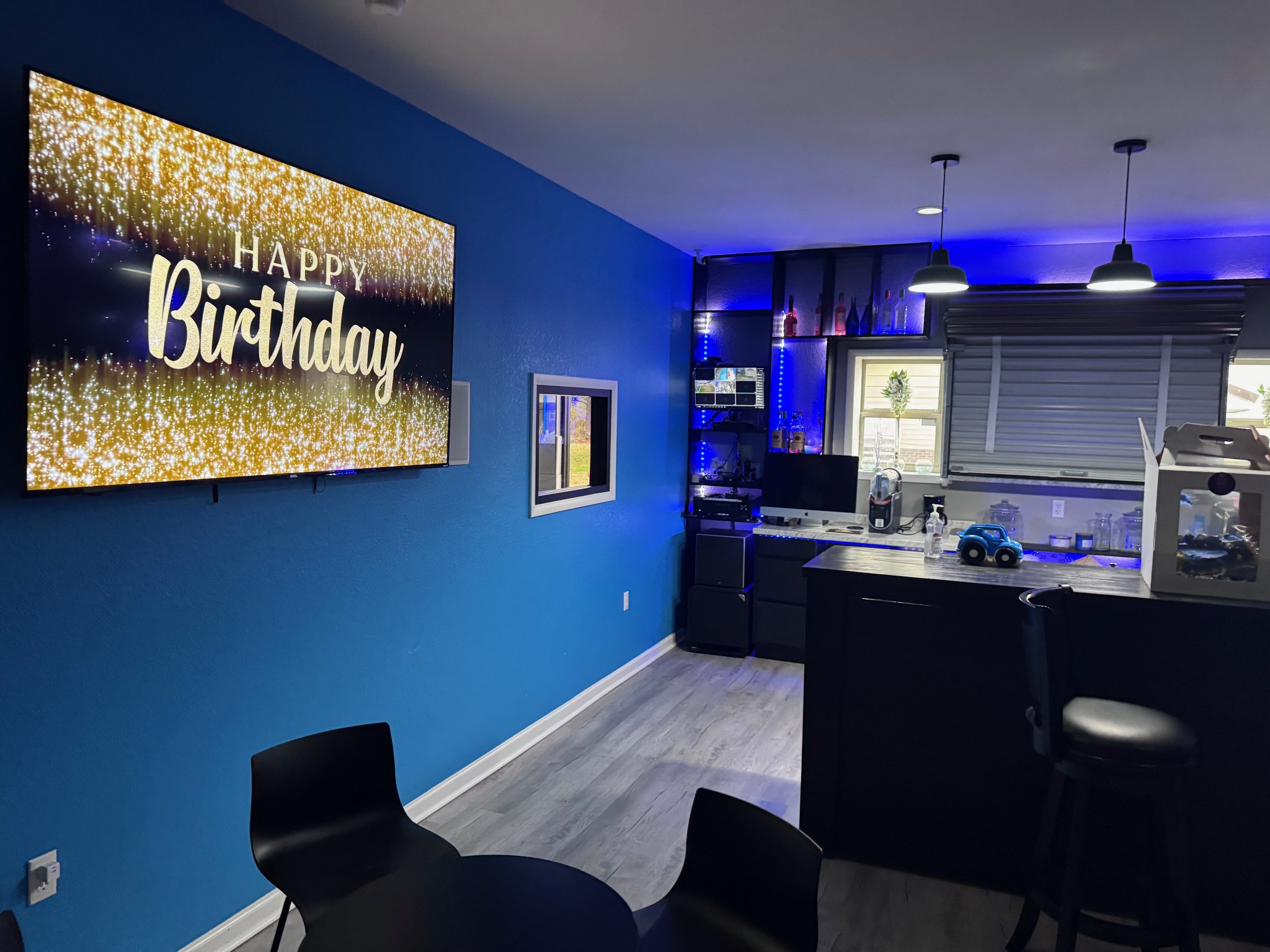 Living room with a large television displaying 'Happy Birthday' with a golden glitter background, a blue accent wall, a small window, a desk with a computer and various items, bar stools, a kitchen counter with toys and a cake box, and blue LED light