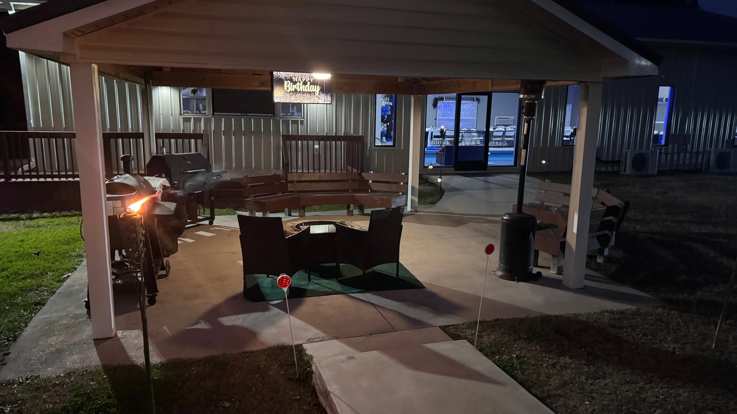 A covered outdoor patio at night with a barbecue grill, sitting area with a table and chairs, benches, two patriotic yard signs, and a 