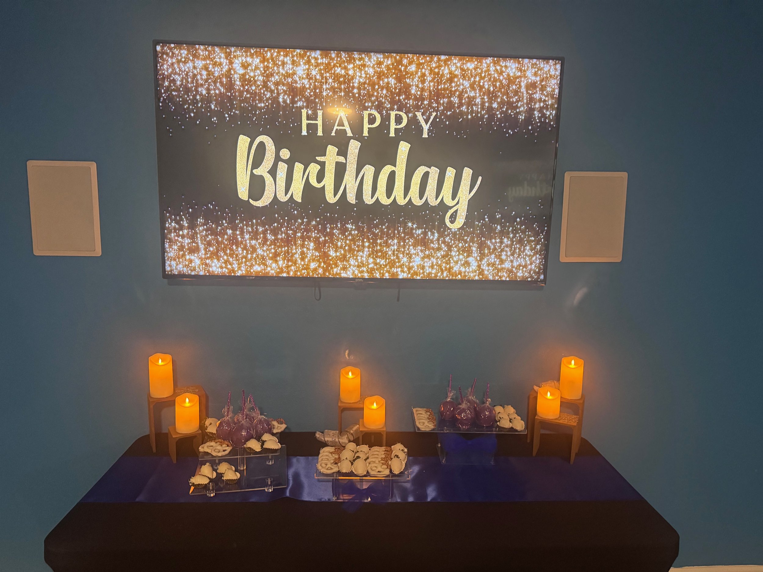 A birthday celebration setup with a large screen displaying 'Happy Birthday' on a glittery background, surrounded by candles, flowers, and treats on a table.