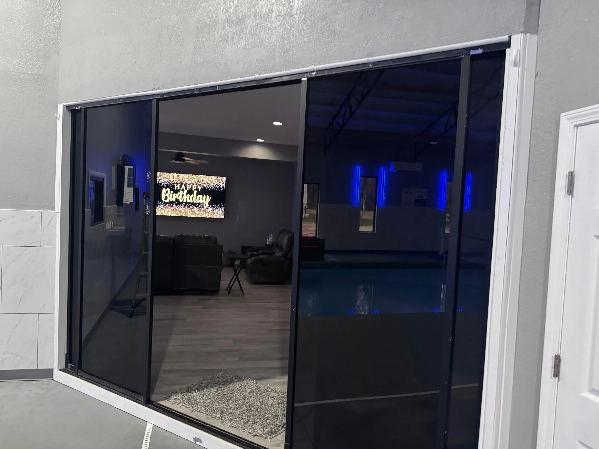 Indoor space with a large sliding glass door, living room with black chairs, sofa, and a TV screen displaying 'HAPPY Birthday' in the background. Blue lighting accents are visible inside.