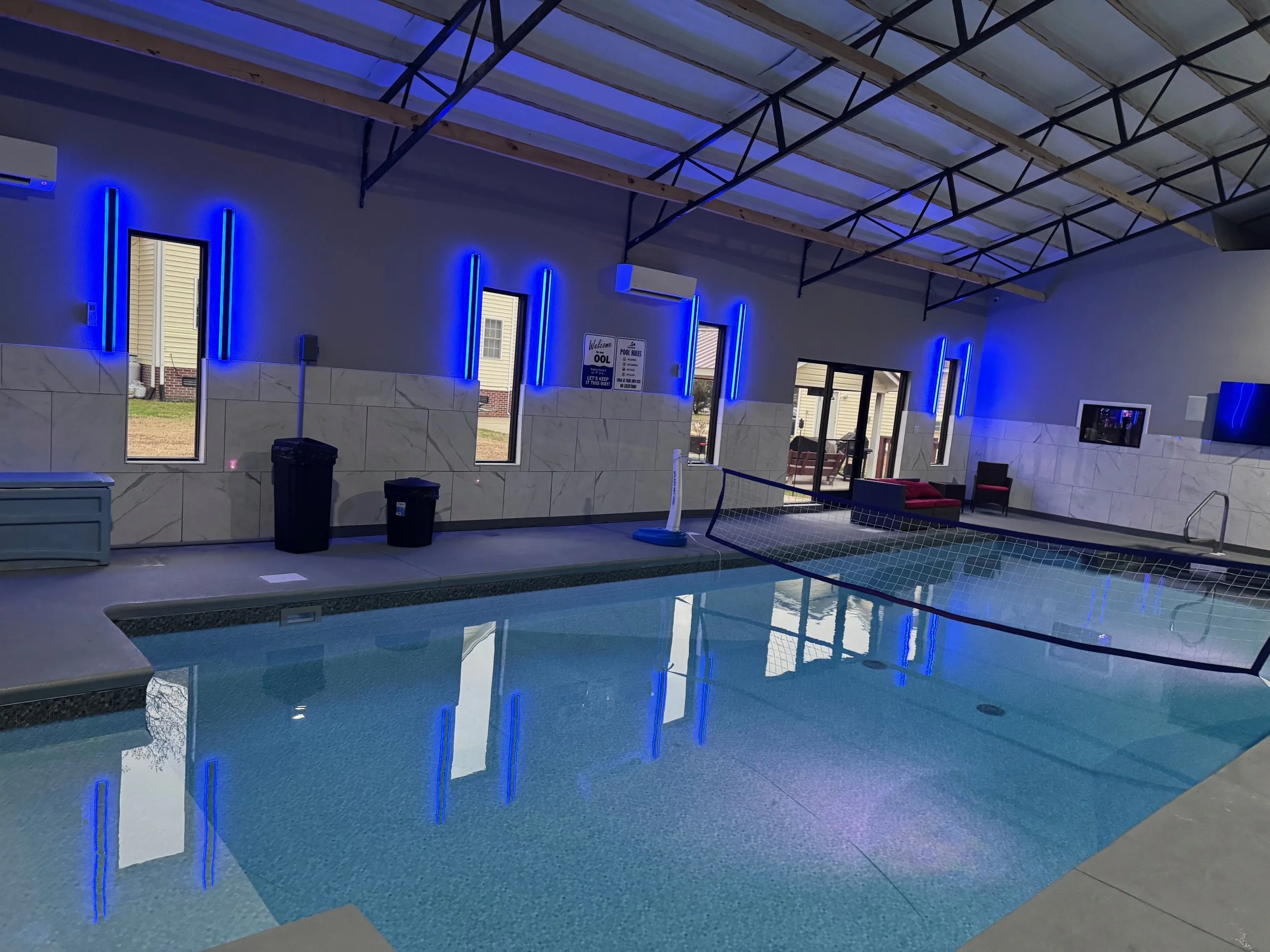 Indoor pool area with neon blue lighting, tiled walls, and lounge chairs, featuring a pool table net and windows.