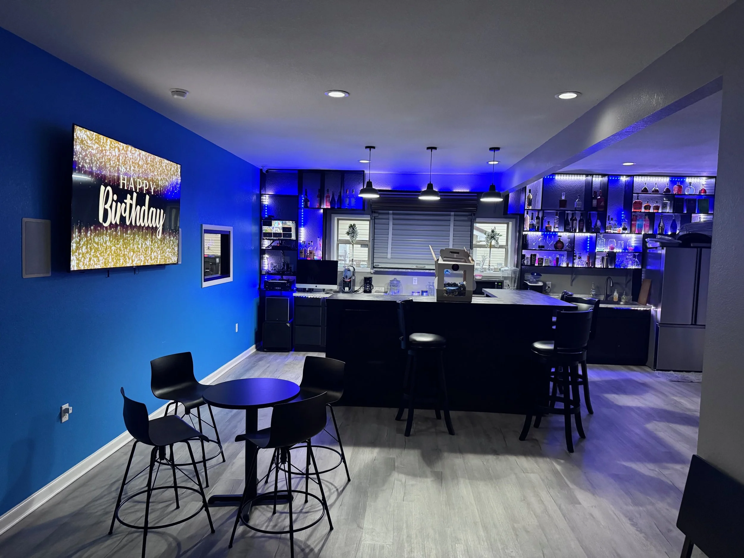Home bar area with blue walls, a flat-screen TV displaying birthday wishes, a round table with four chairs, and a bar counter with barstools. Shelves behind the bar are illuminated in blue with bottles and glasses.
