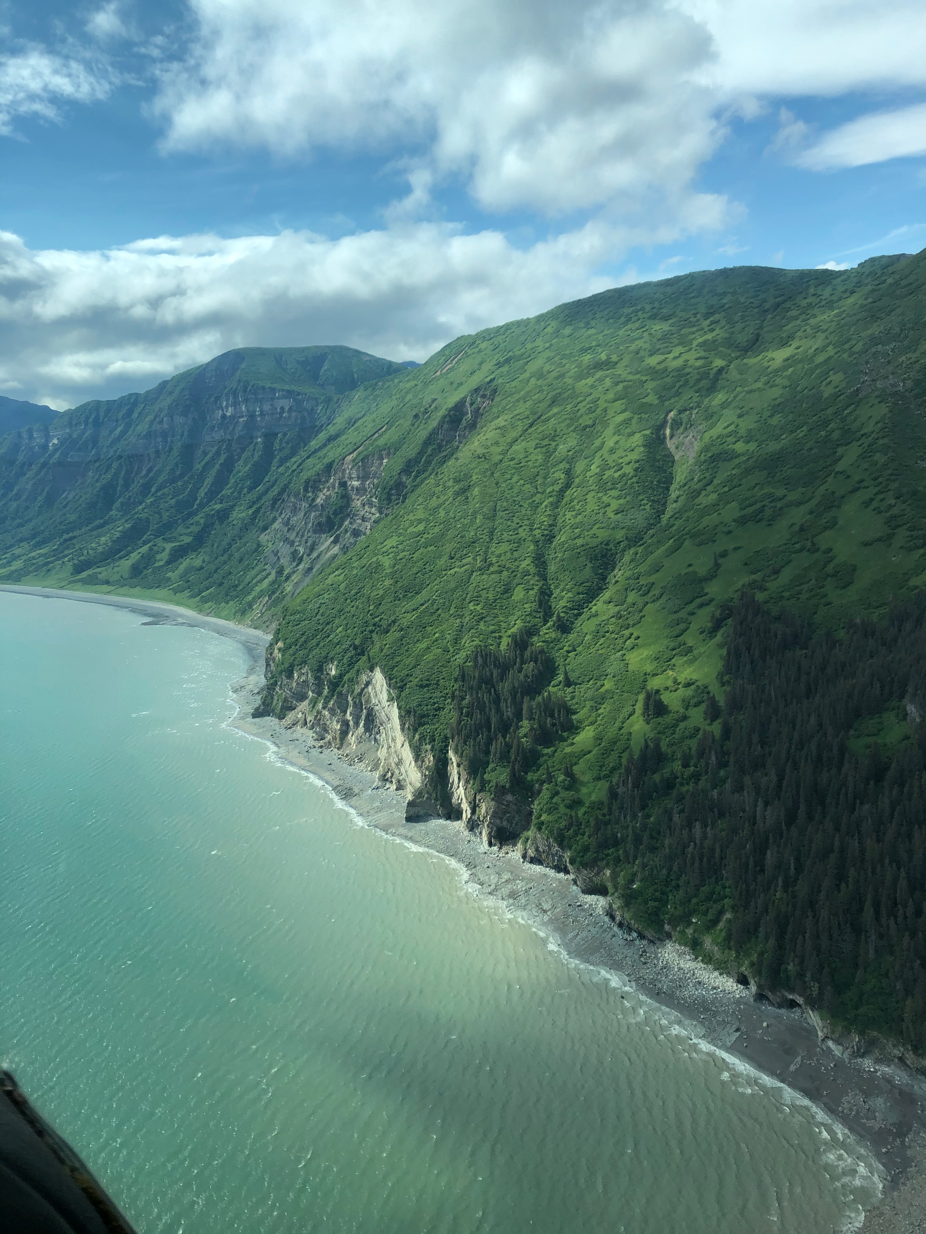 Aerial view of a green mountainous coastline adjacent to a calm body of water, with partly cloudy skies overhead.