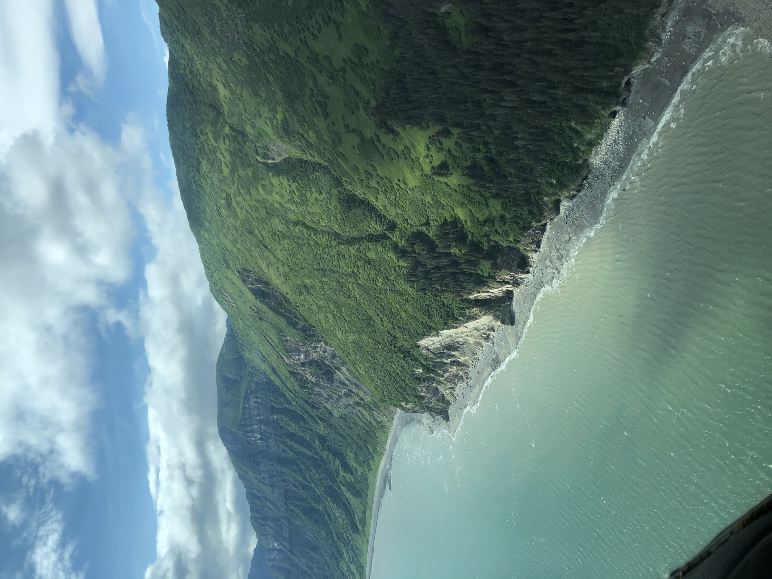 Aerial view of a green mountainous coastline adjacent to a calm body of water, with partly cloudy skies overhead.
