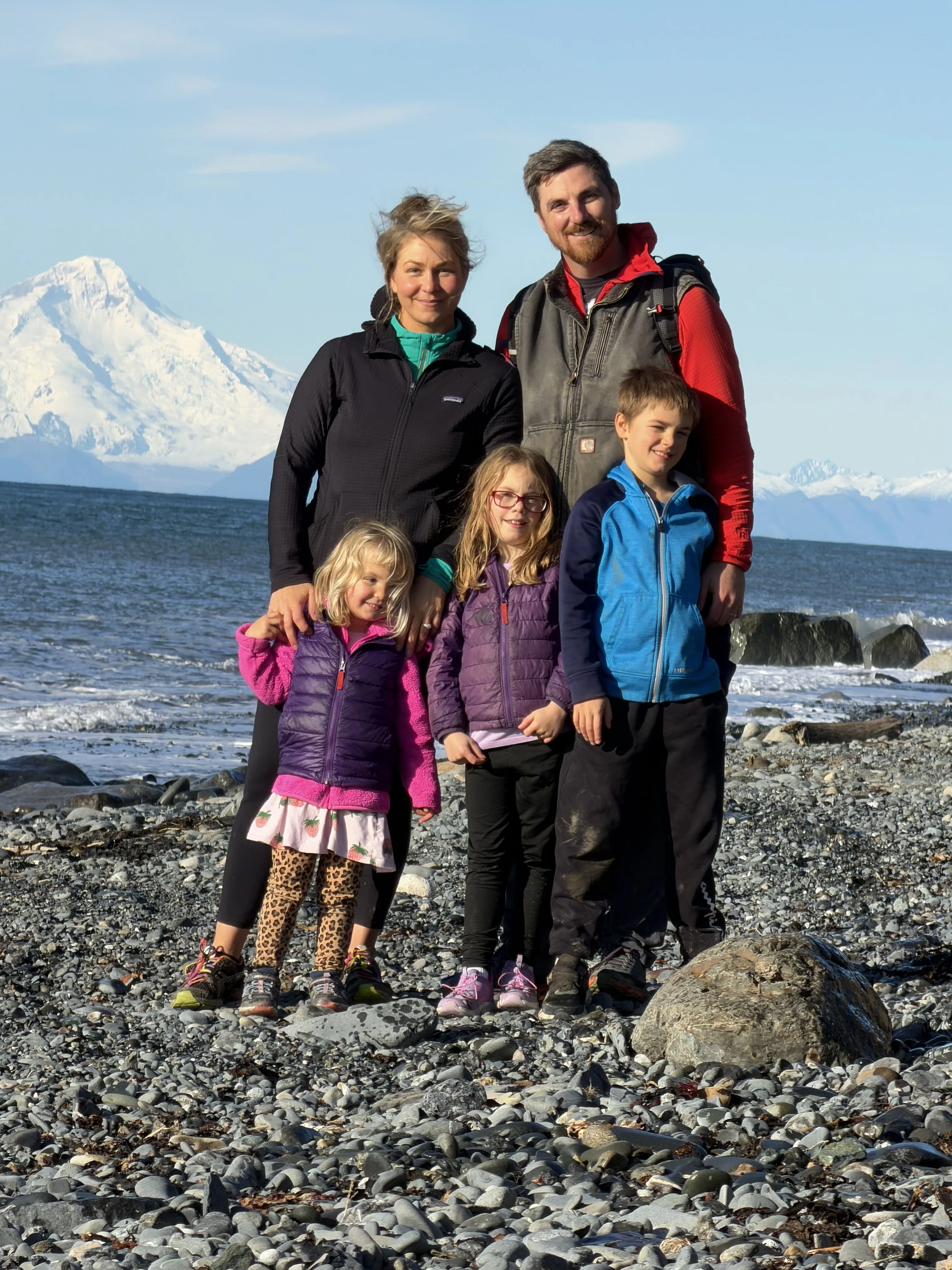 Family of six in outdoor clothing standing on rocky beach with snow-covered mountain and water in the background.