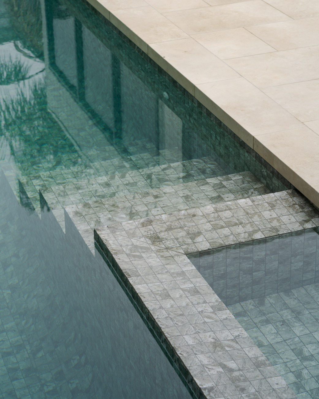 Close-up of a swimming pool with clear water, showing the tiled pool floor and the pool's edge next to a beige stone deck.