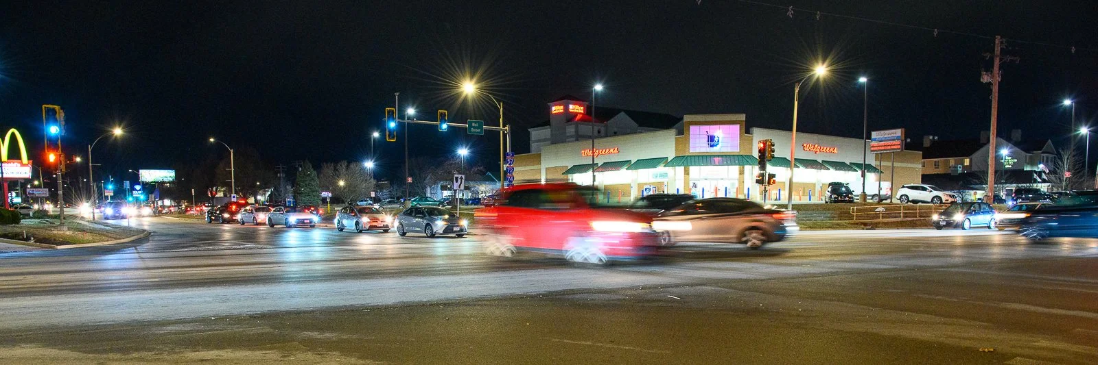 Night scene of a busy intersection with moving cars, traffic lights, and a Walgreens store in the background, illuminated by streetlights.