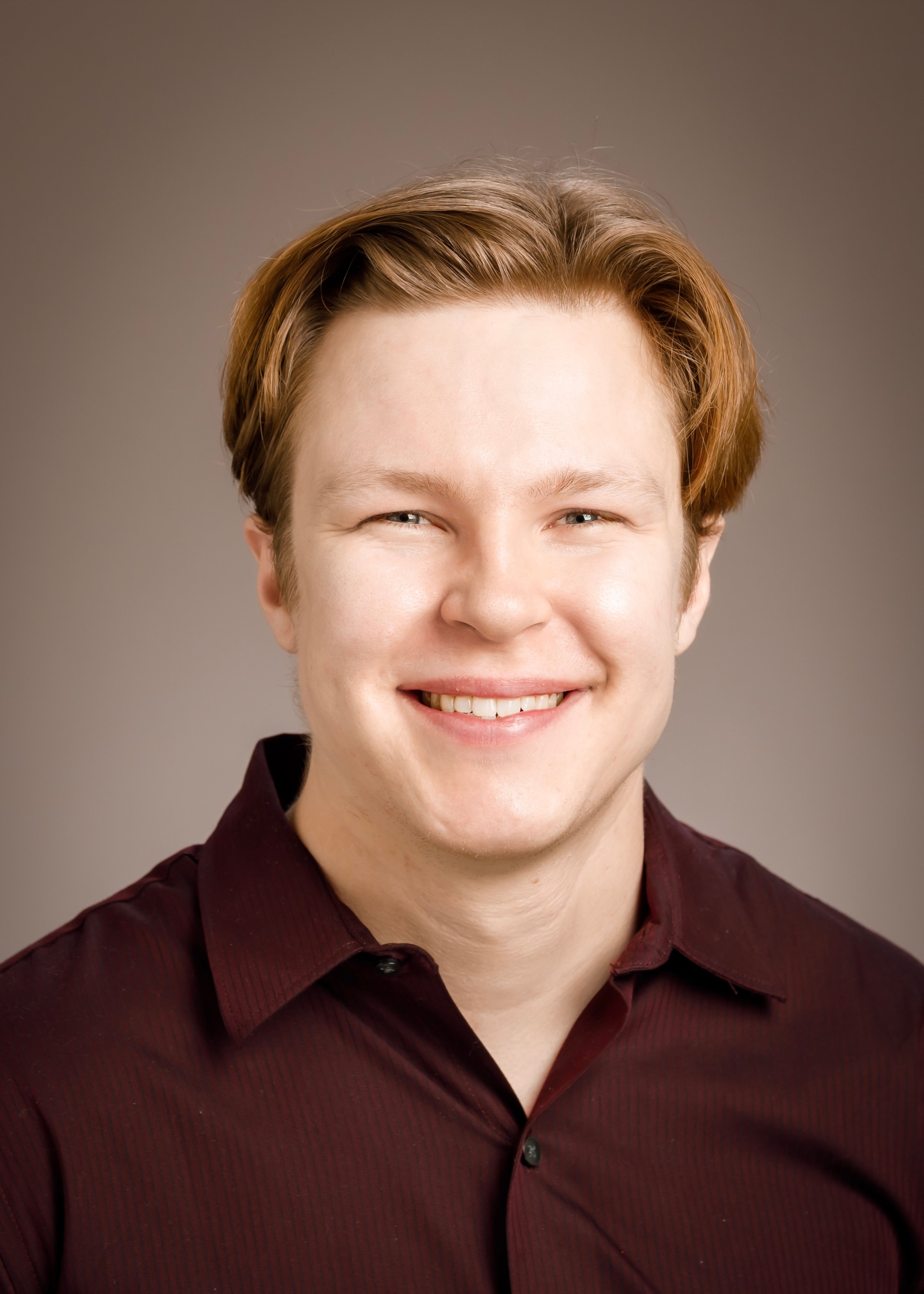 Close-up of a young man with short, wavy brown hair, smiling, wearing a dark maroon button-up shirt, against a neutral background.