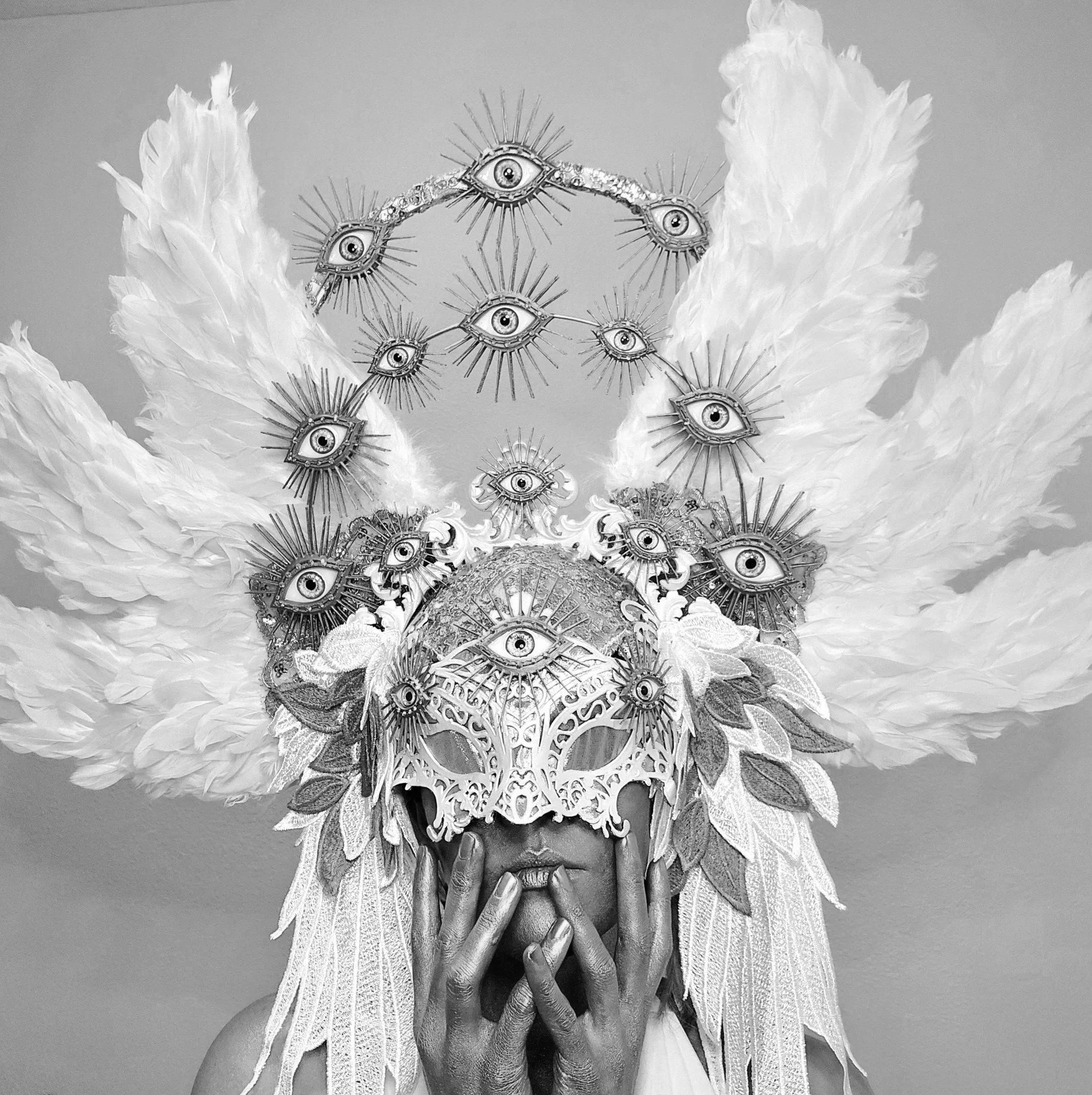 A black-and-white photo of a person with elaborate headwear decorated with many eye symbols, surrounded by large feathers and lace, with their hand near their chin.