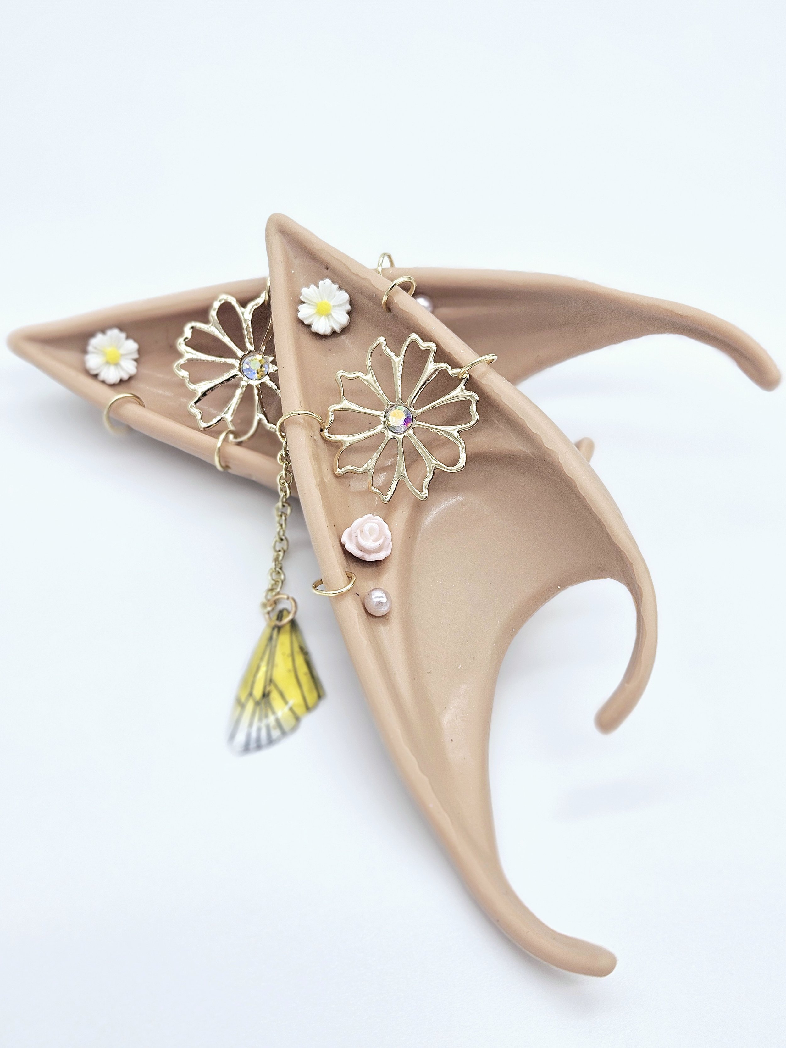 A pink ceramic dish shaped like a human ear with jewelry and decorative items, including a daisy, a pink rose, a yellow and black bee, and gold jewelry, arranged inside and on the ear.