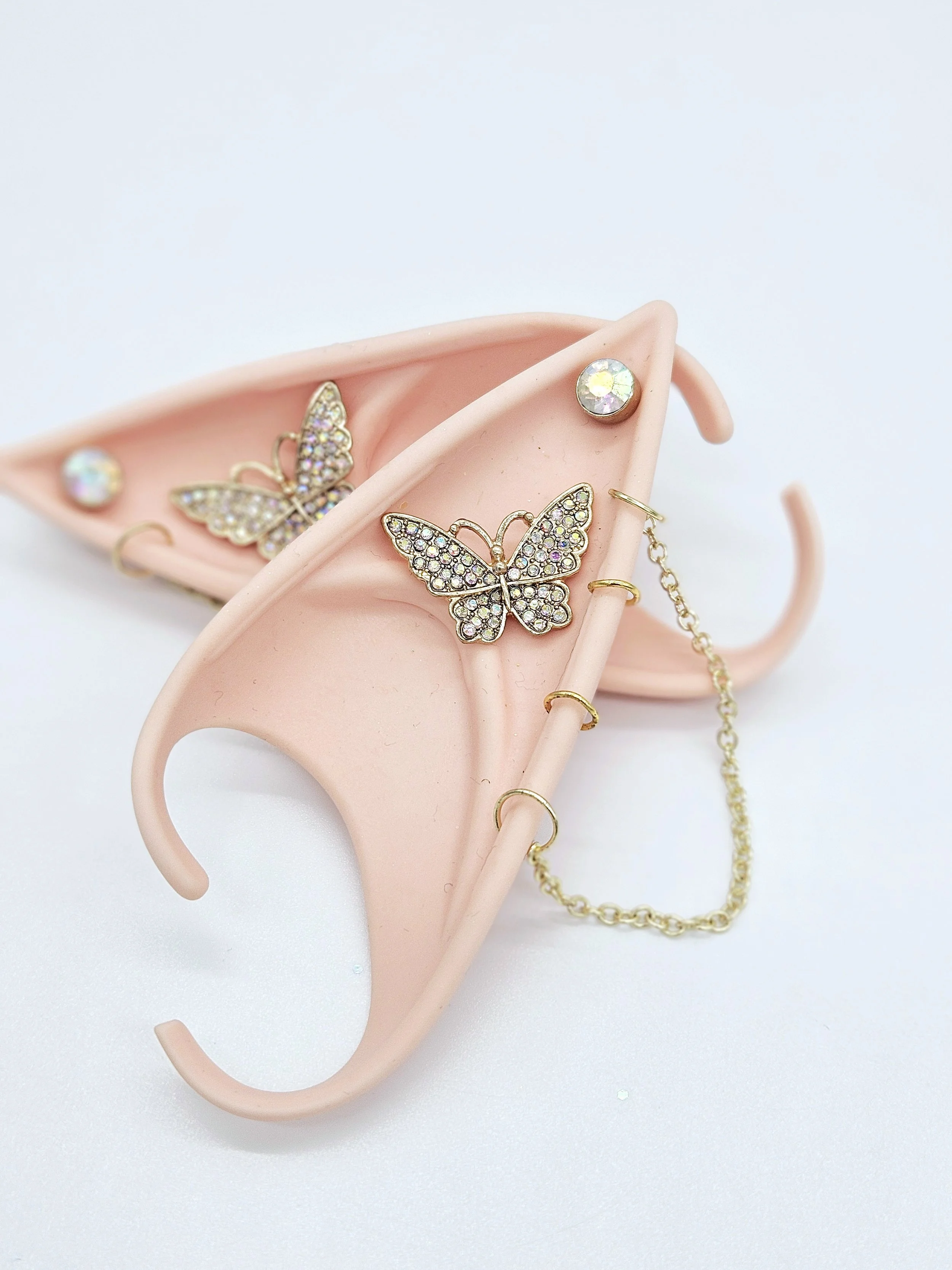 A light pink jewelry holder in the shape of a human ear, holding butterfly-shaped earrings with rhinestones, a round rhinestone stud earring, and a gold chain necklace.