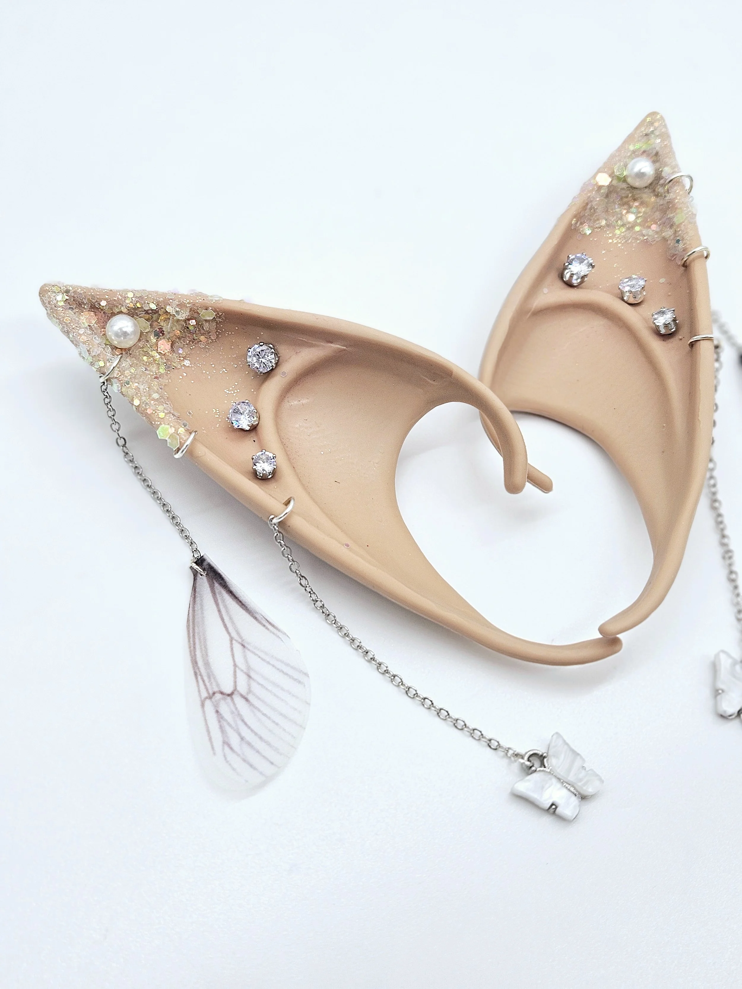 A beige mannequin ear adorned with sparkling earrings resembling a dragonfly, decorated with glitter, pearls, and rhinestones, with matching jewelry displayed on a white surface.