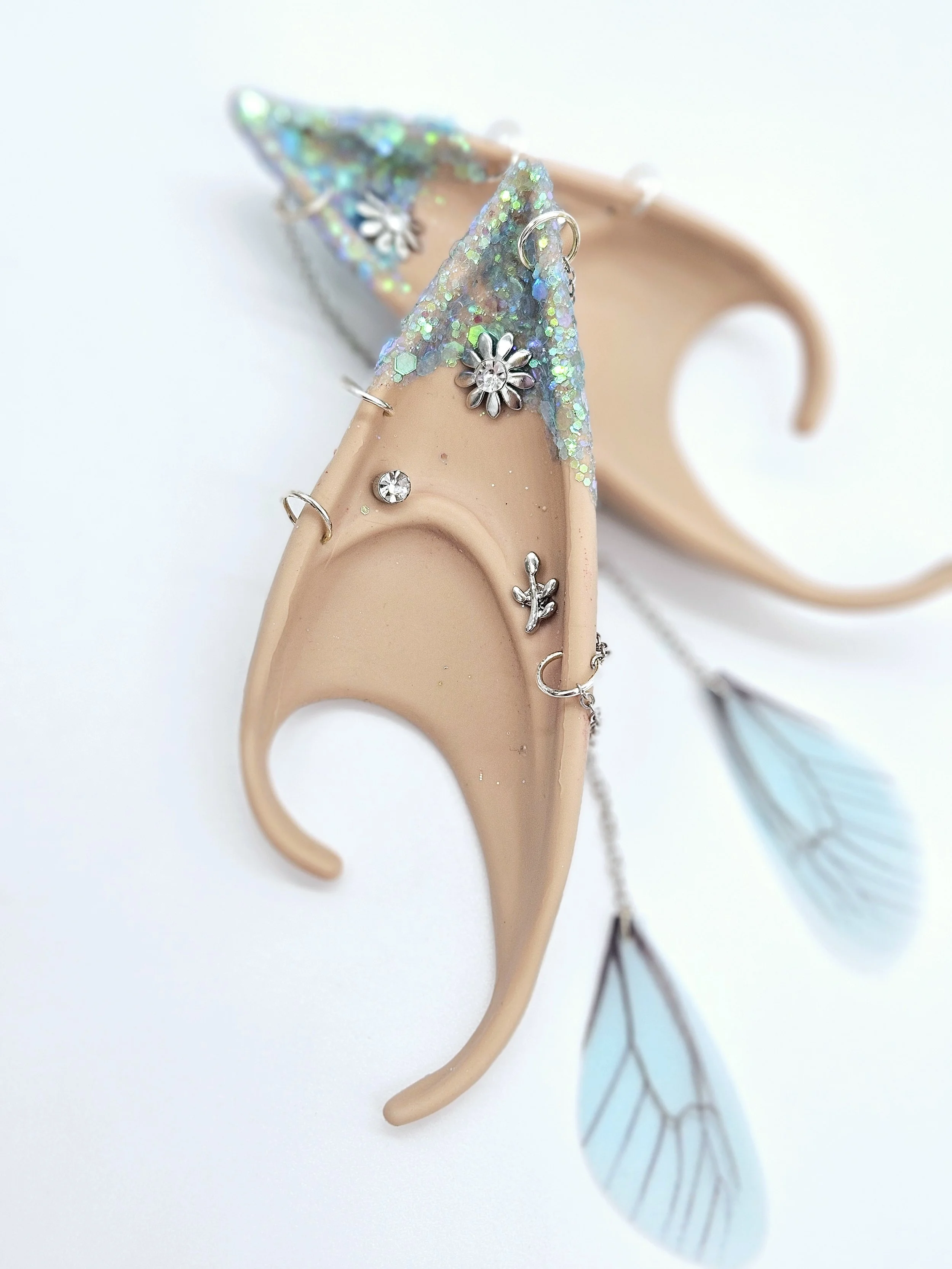 Close-up of a beige shoe decorated with silver flower charms, glitter, and rhinestones, with a blue leaf in the background.