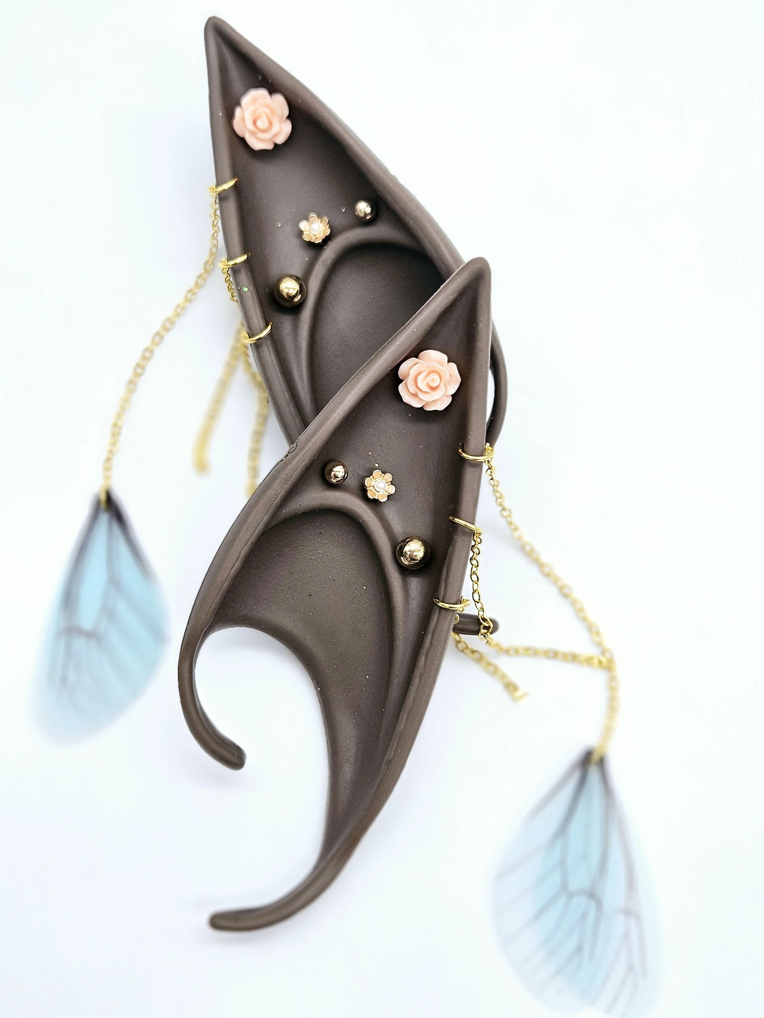 Decorative pair of eyeglasses with brown frames, pink and gold floral embellishments, gold chains, and blue feathers attached to the sides, set against a white background.