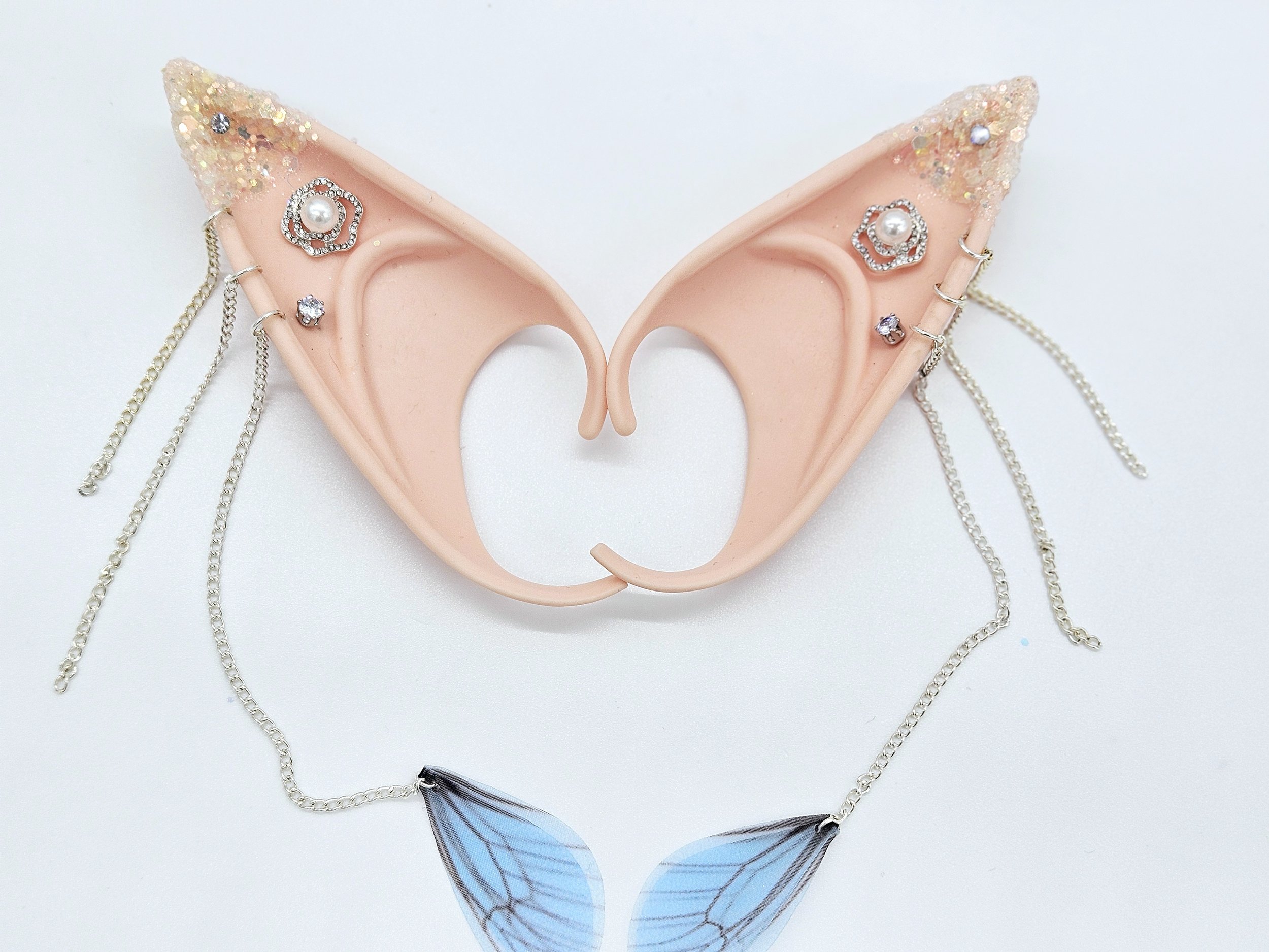 Pink silicone fairy or elf ears adorned with jewelry, rhinestones, pearls, and chains, with a blue butterfly decoration attached at the bottom.
