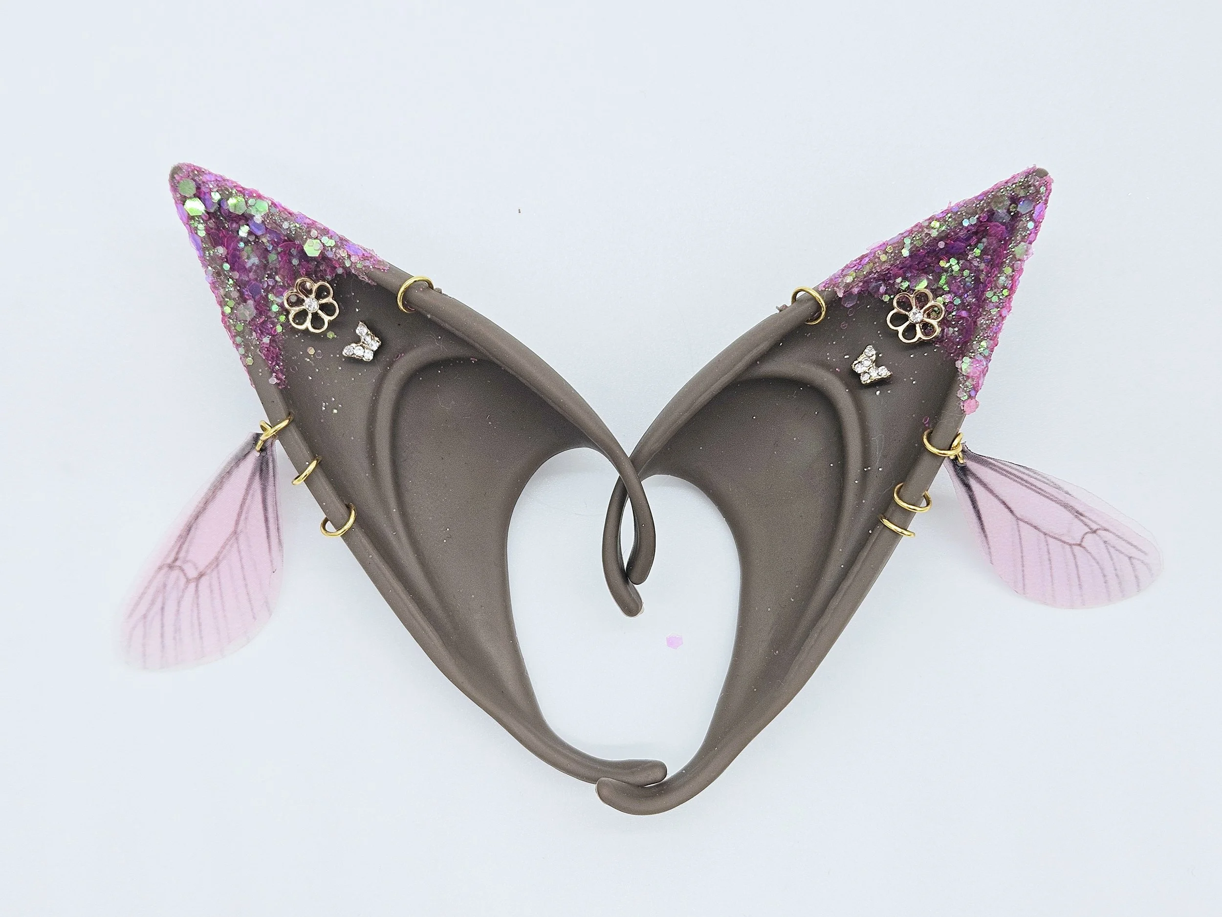 Close-up of a decorative hair accessory resembling fairy or elf ears with pink and purple glitter, small flower and butterfly embellishments, and translucent pink wings.