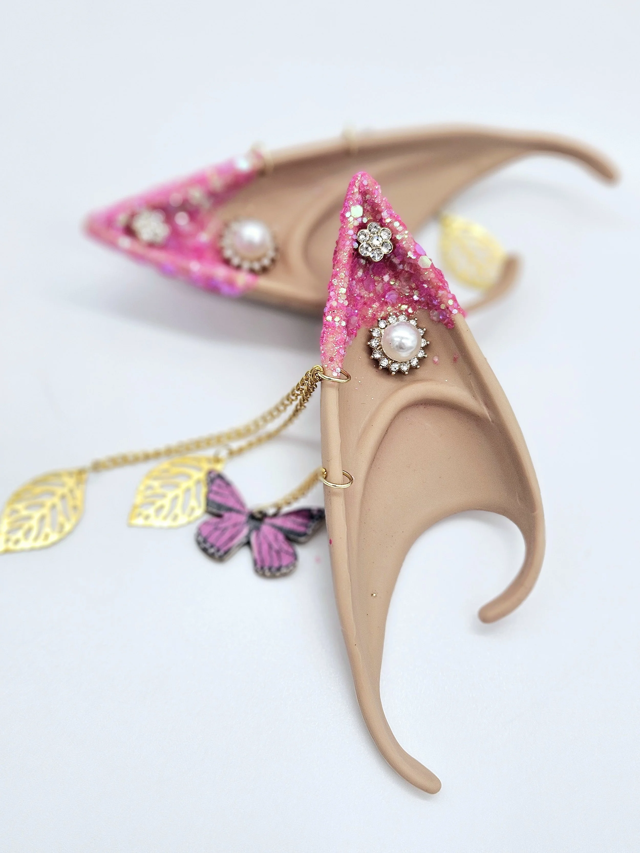 A pink, glittery hair clip decorated with rhinestones and pearls is placed on a beige plastic jewelry display stand shaped like a hand, with a butterfly jewelry piece and gold earrings also visible in the background on a white surface.