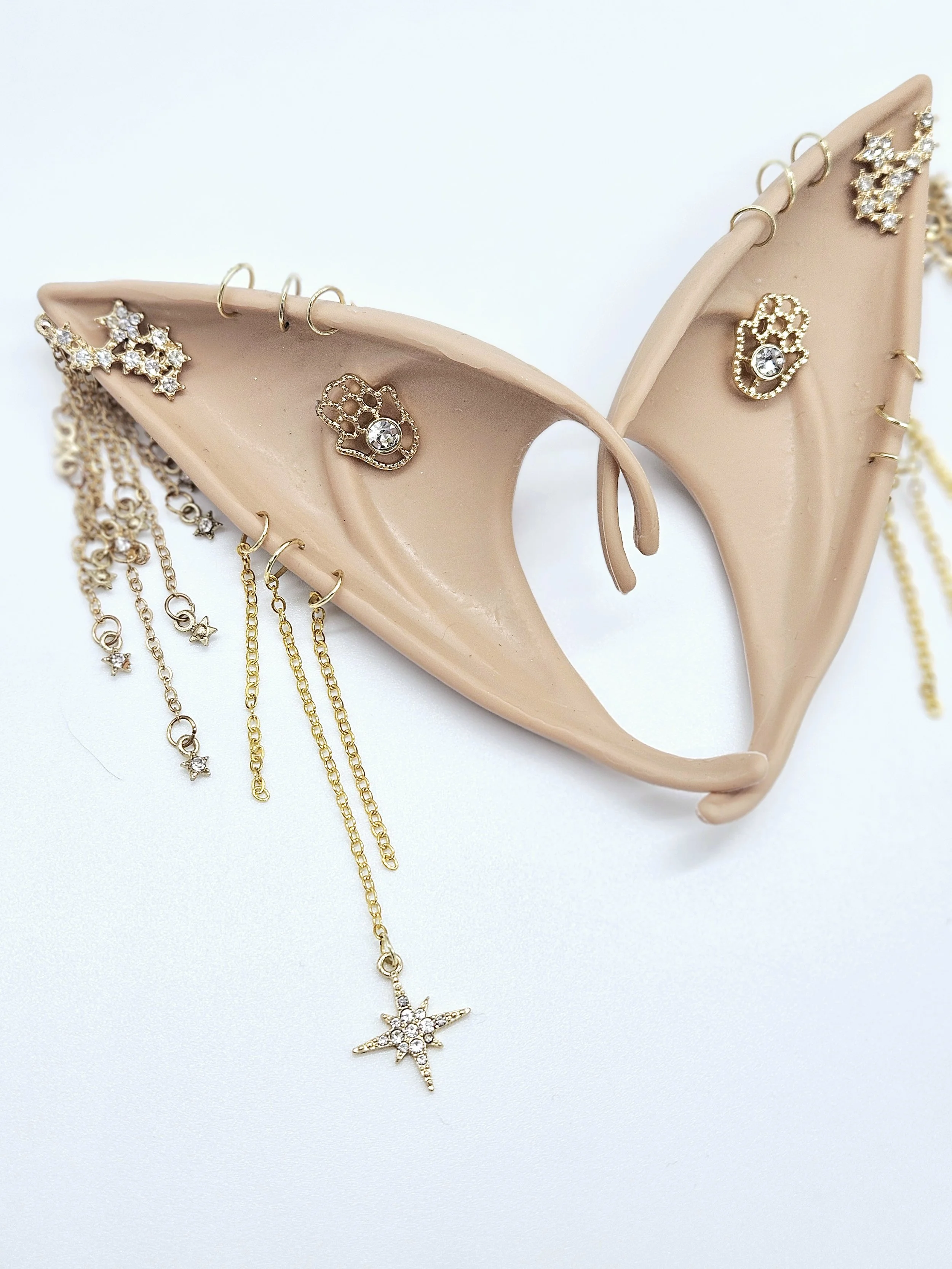 A beige high heel shoe decorated with gold jewelry, including star-shaped, heart-shaped, and starburst-shaped earrings, and hanging necklaces with star and gemstone pendants.