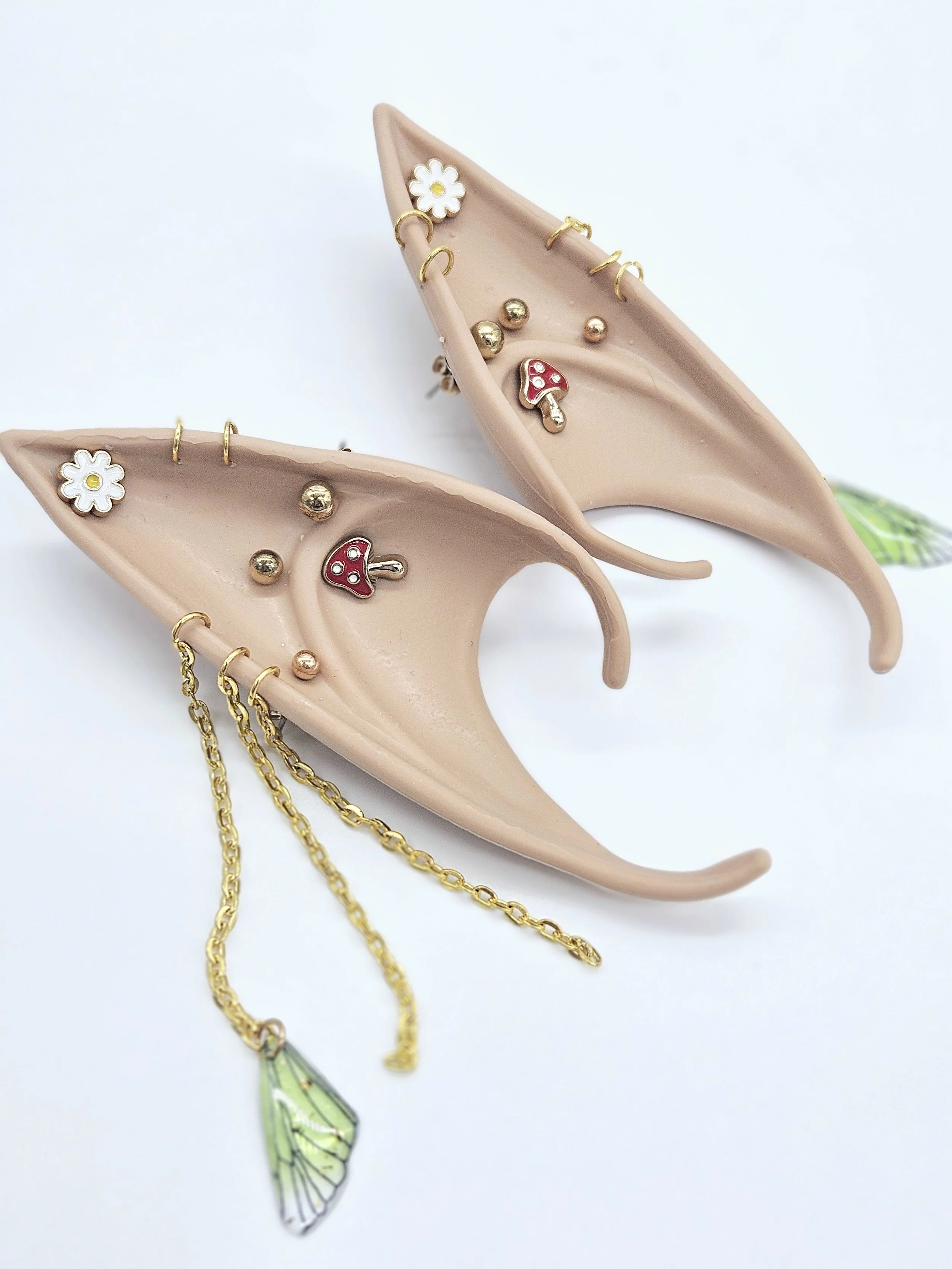 Two beige leaf-shaped dishes decorated with small jewelry charms including white daisy, red and white mushroom, gold and silver balls, and a green and black butterfly pendant, on a white background.
