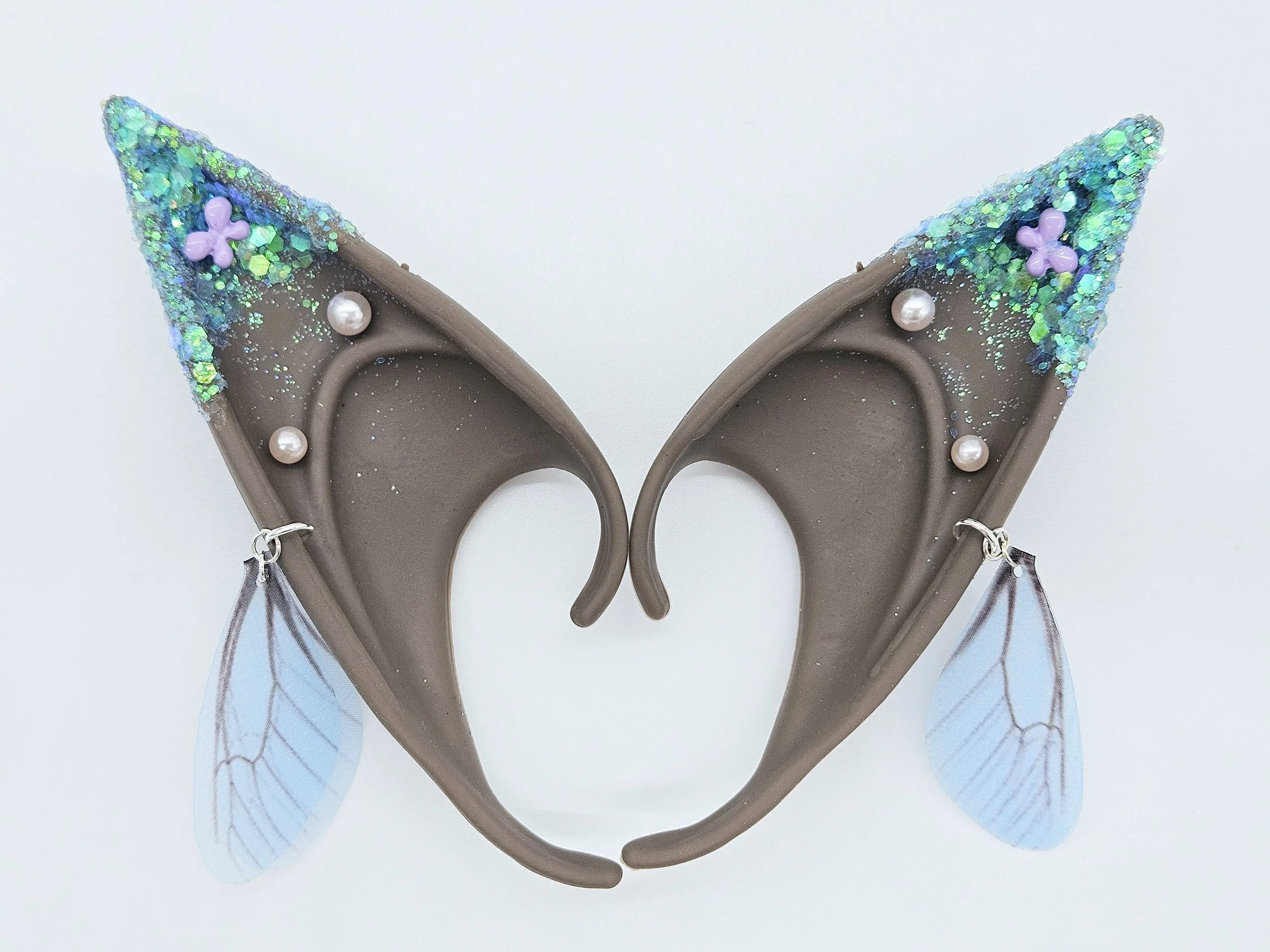 A pair of butterfly-shaped hair clips decorated with glitter, small pearl accents, and purple flower decorations, featuring light blue and silver wings with delicate veining.