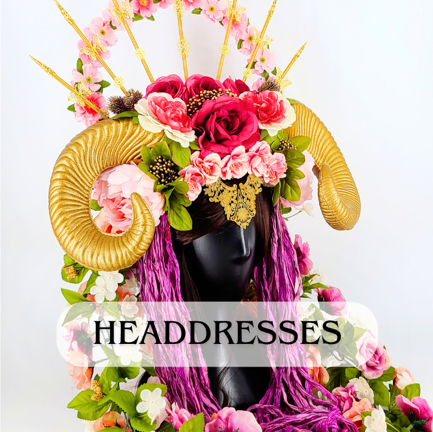 A decorative headpiece featuring large gold ram horns, pink and red flowers, green leaves, and purple yarn hanging down, displayed on a black mannequin head. The word 'HEADDRESSES' is written across the image.