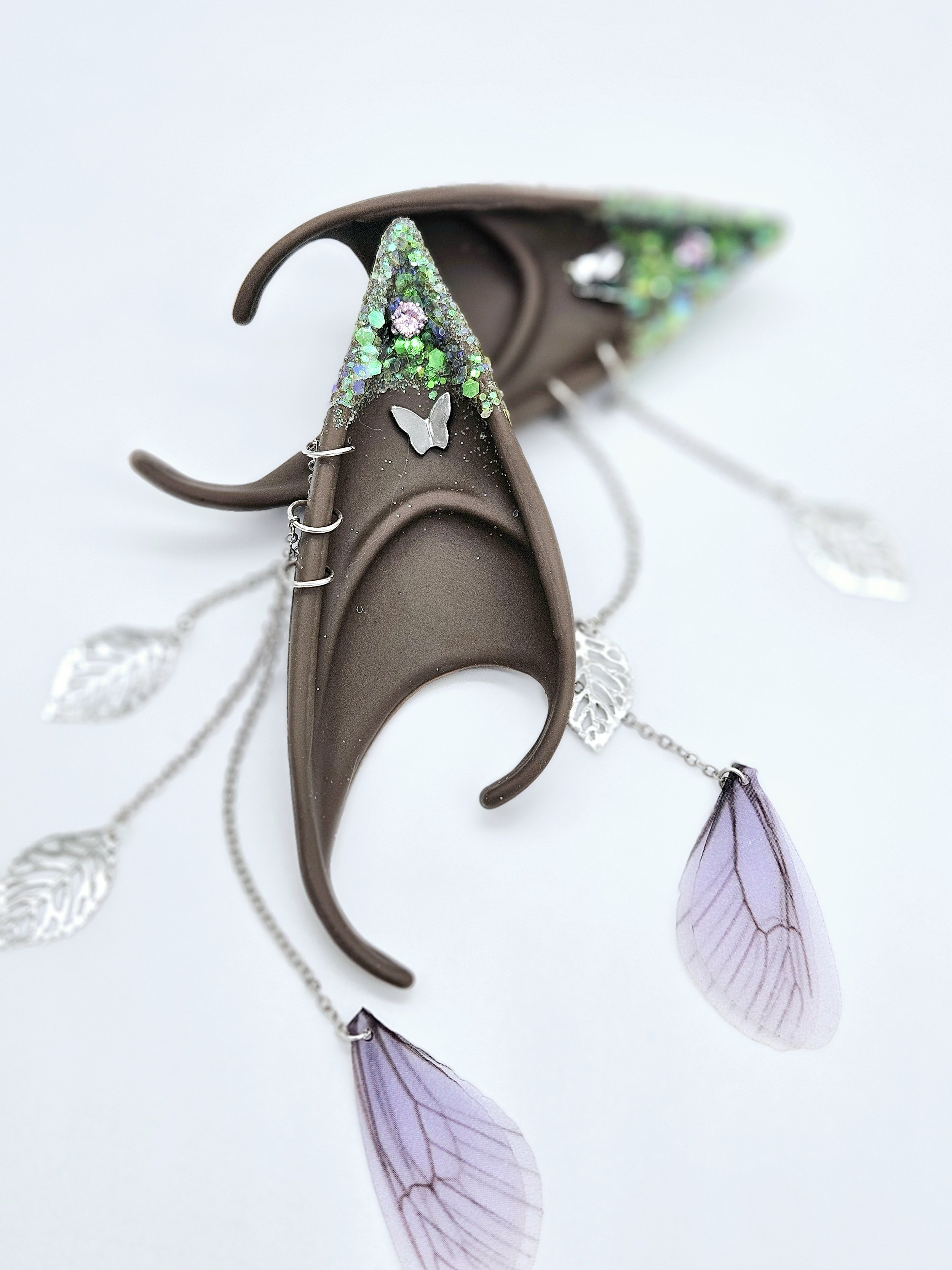 Decorative hair clip shaped like a butterfly with glitter, attached to a brown headband with purple transparent wings and silver leaf charms, set against a white background.