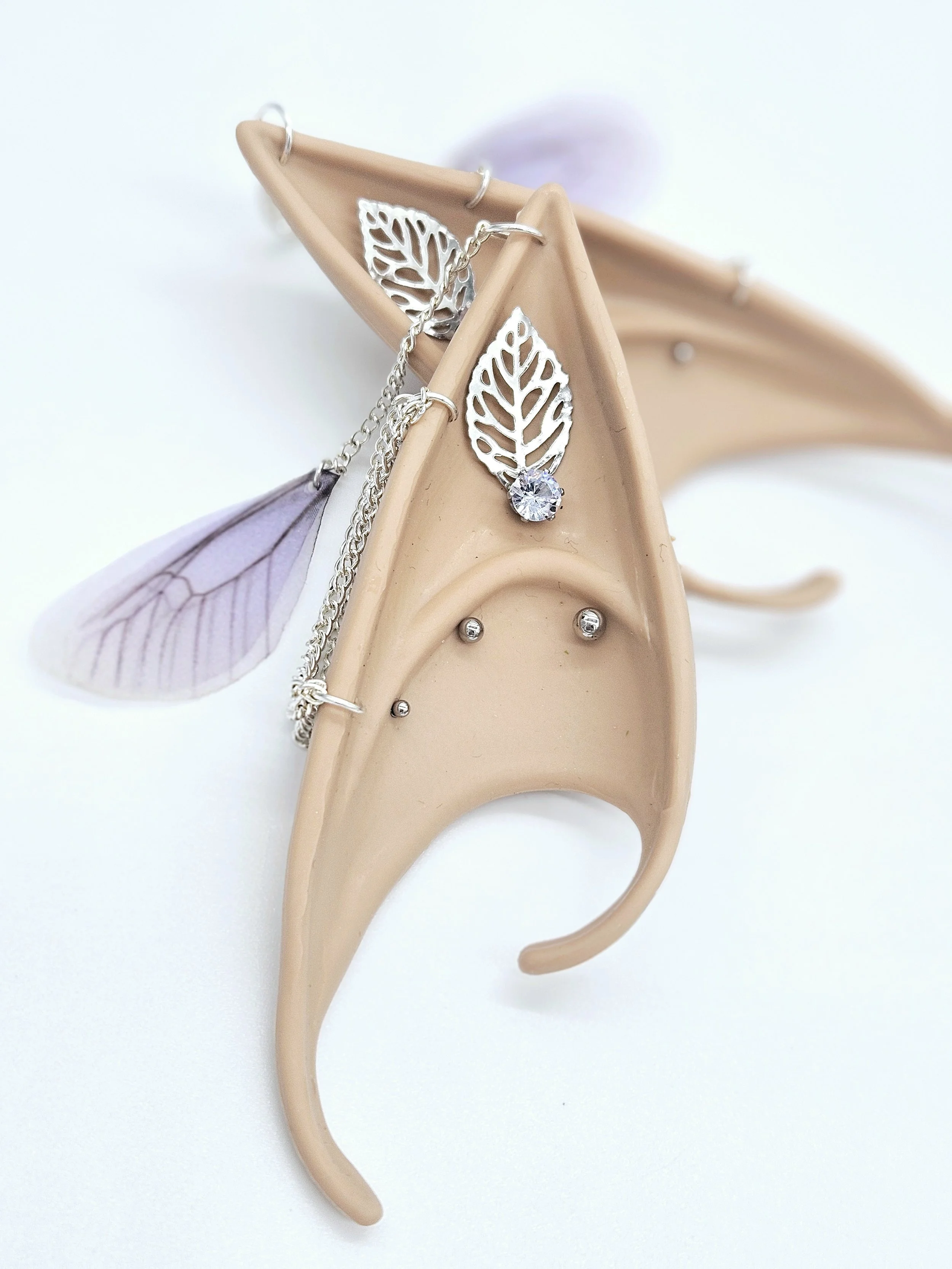 A beige plastic or ceramic decorative earring holder shaped like a human ear with small silver earrings, a leaf-shaped pendant with a rhinestone, and a silver chain with a leaf-shaped charm hanging from a jewelry display.