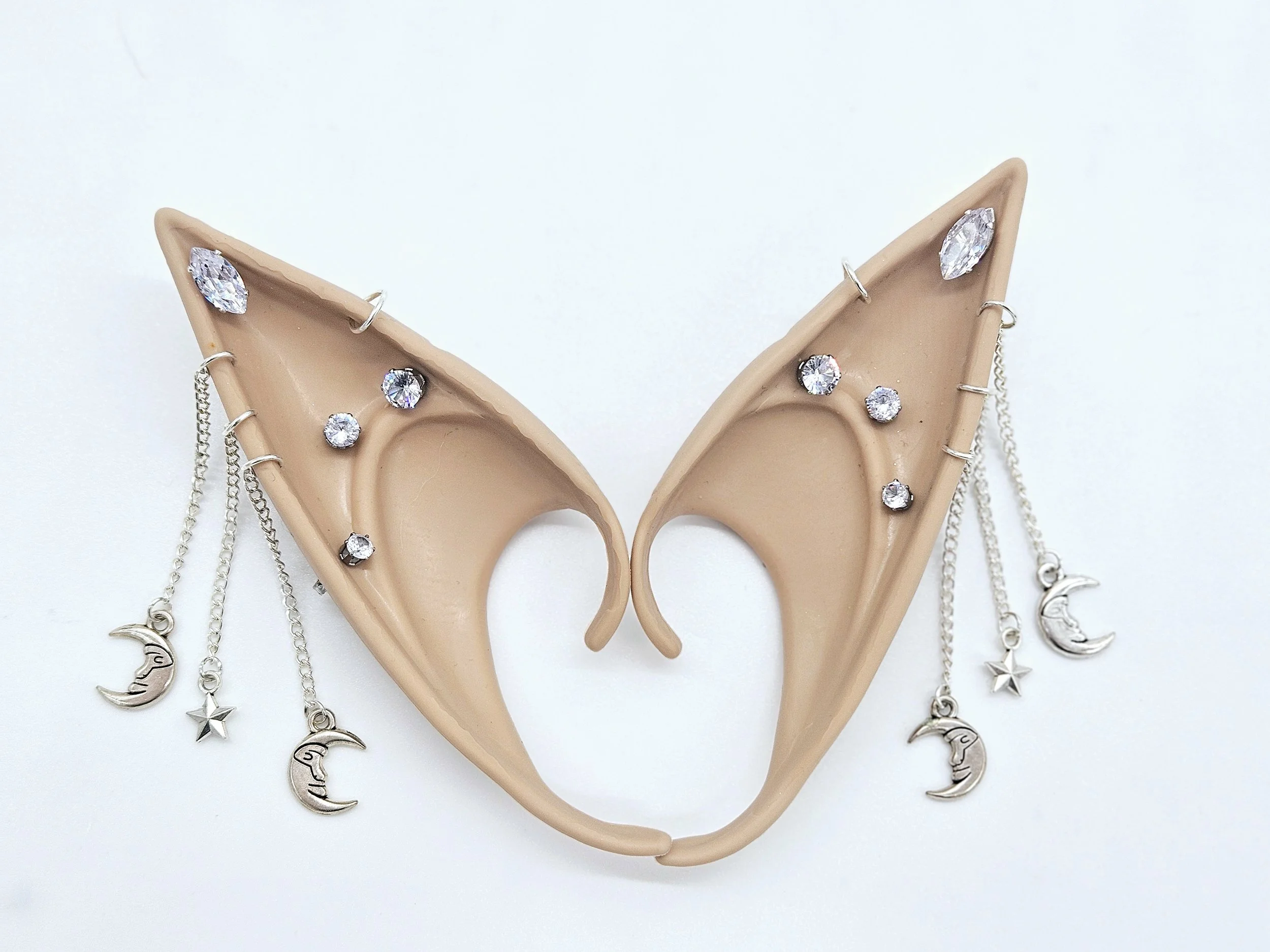 A pair of beige mannequin ears decorated with rhinestone earrings and necklaces featuring moon and star charms, set against a plain white background.